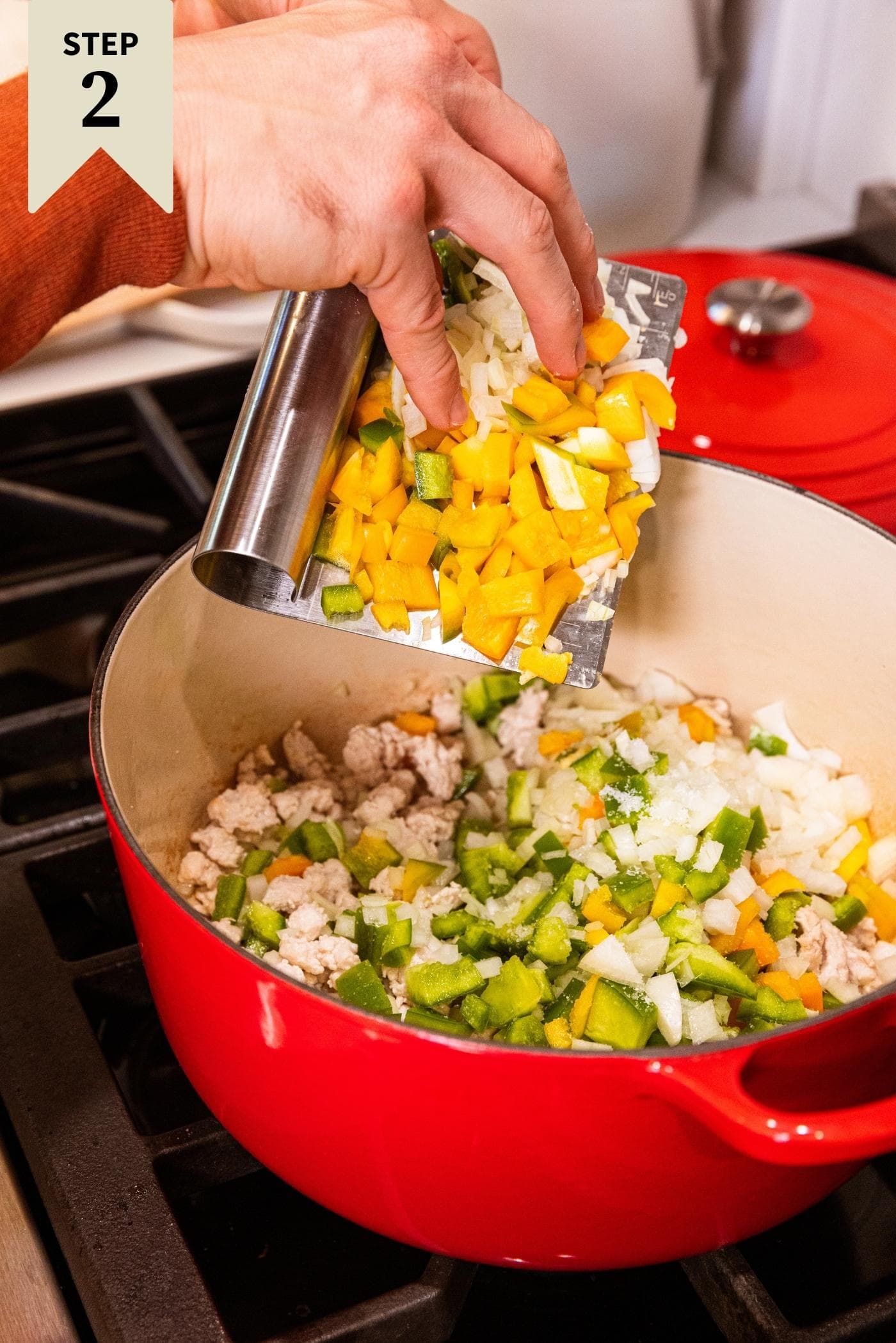 Metal bench scraper dropping chopped yellow and green bell pepper and onion into a red Dutch oven with ground turkey in the bottom already on stovetop.