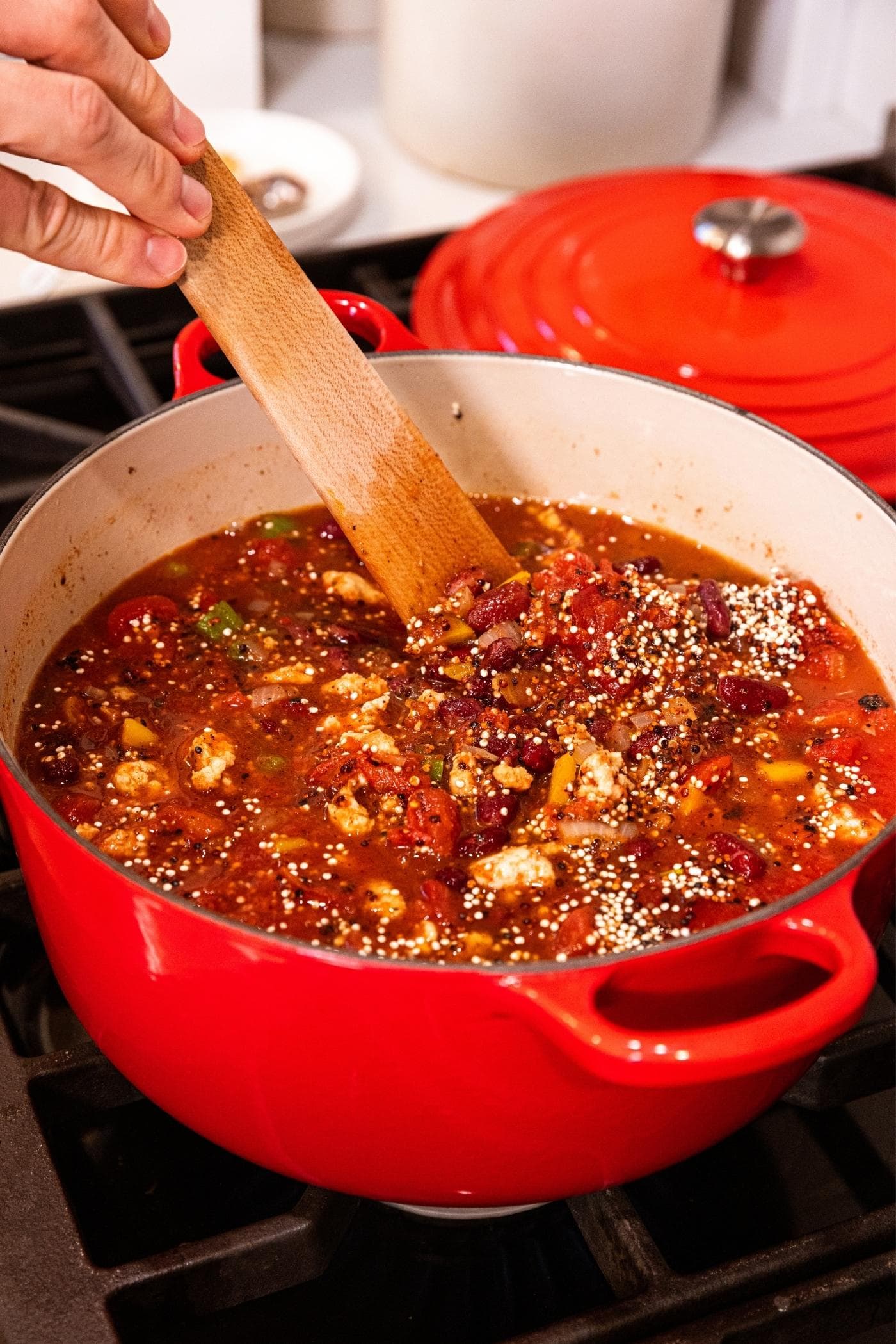 Hand stirring together stoup with quinoa sitting on top before being stirred into red colored soup in Dutch oven on stovetop.