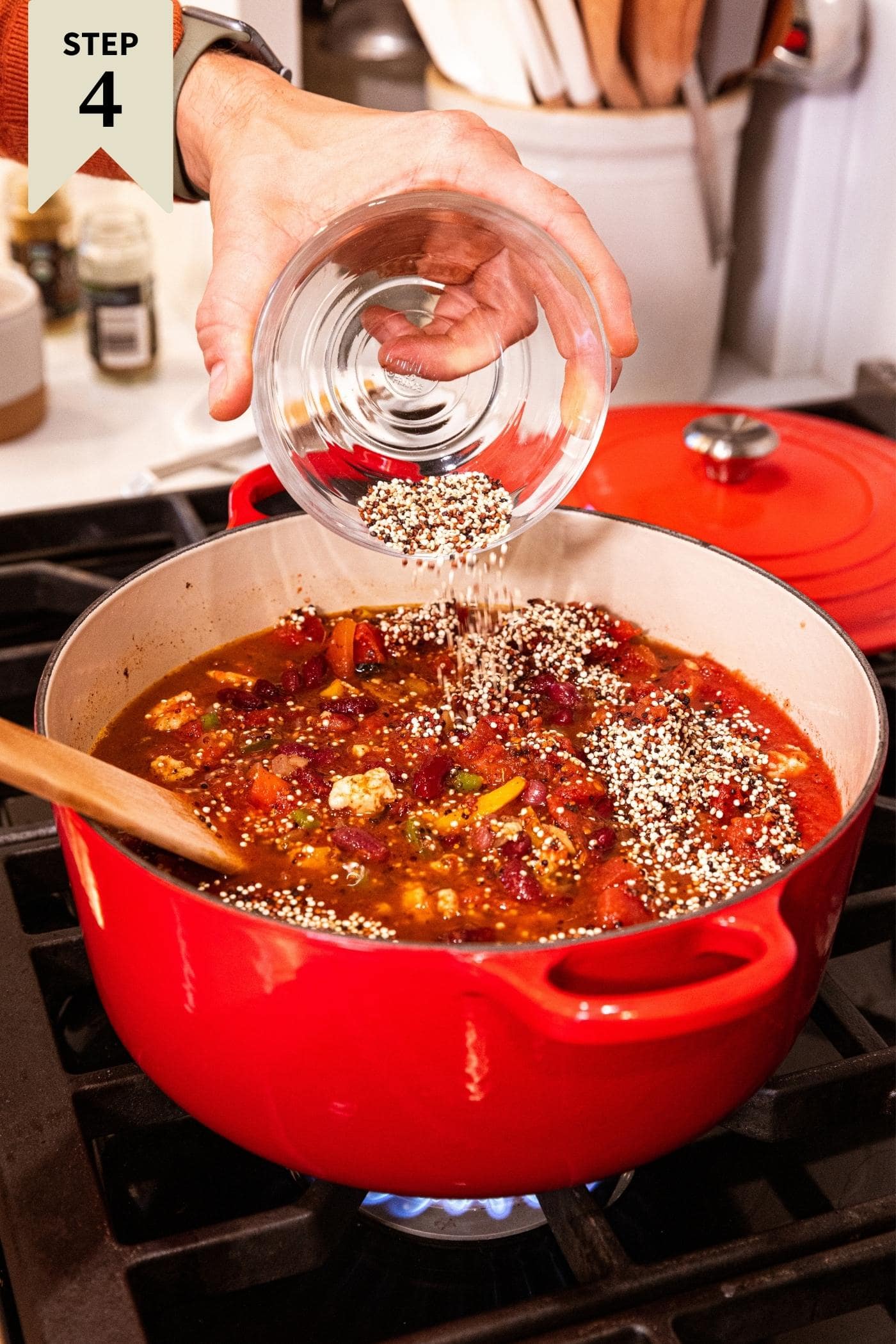 Hand pouring quinoa out of small glass bowl on top of soup sitting in red Dutch oven on stovetop with wooden spatula resting in bowl.