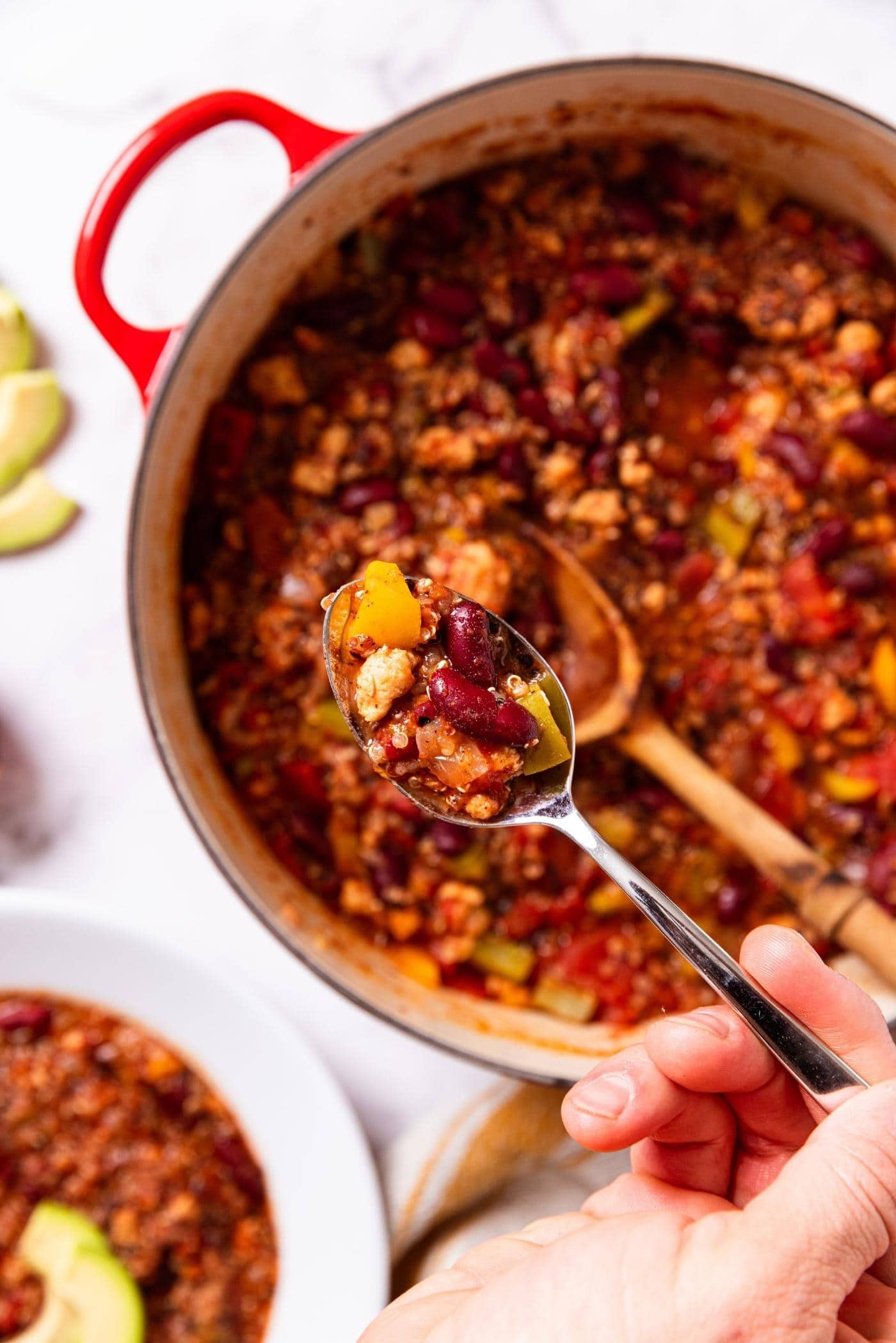 Hand holding a spoon above a red Dutch oven filled with turkey quinoa chili with white bowl to the side also filled with chili.