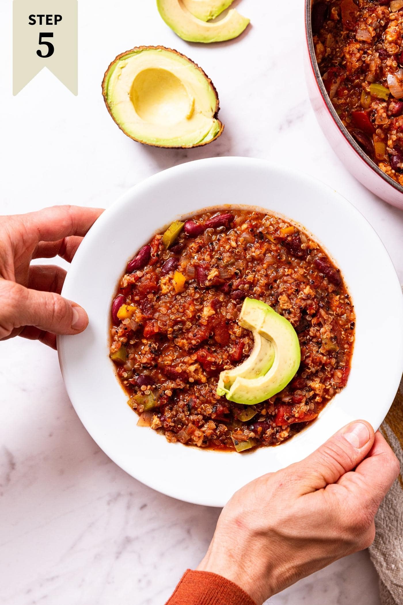 Two hands placing a white bowl filled with turkey quinoa chili topped with slices of avocado onto marble surface with rest of soup to the side.