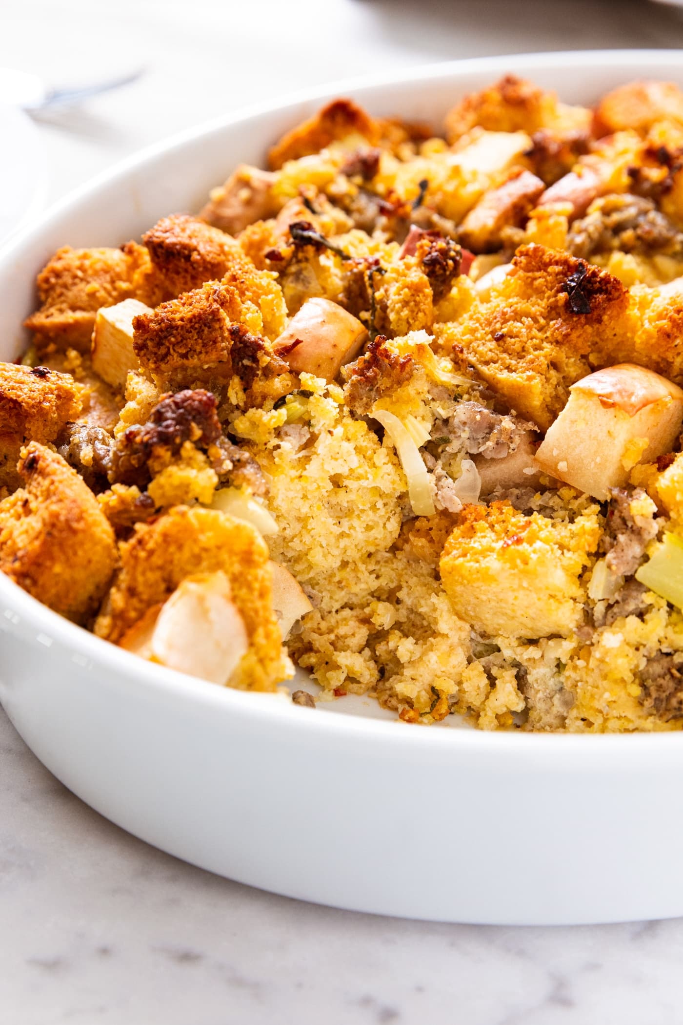 White oval baking dish filled with cornbread stuffing with a scoop taken out showing the cornbread interior with sausage and apple as well.