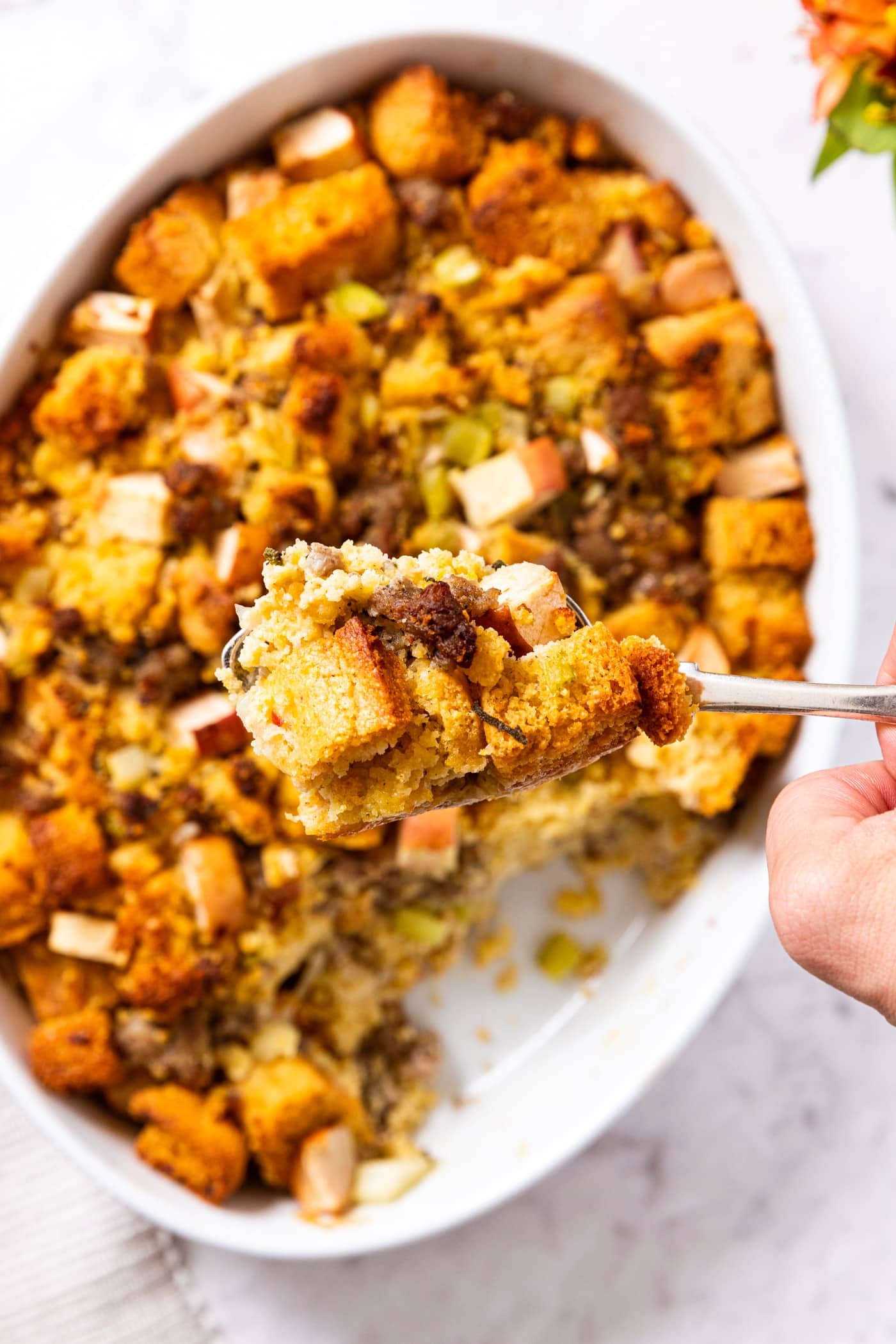 Hand holding a scoop of cornbread stuffing above the rest of the dish filled with rest of the stuffing all on marble surface.