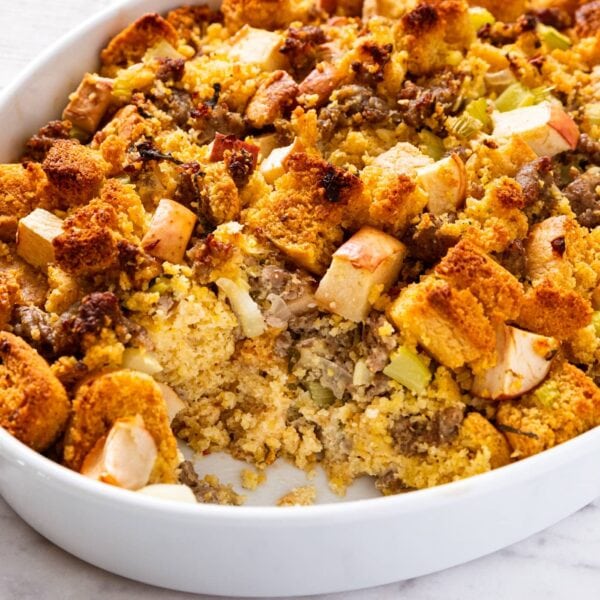White oval baking dish sitting on marble surface after being removed from the oven showing brown crispy top with a scoop missing with interior showing cornbread, apple, and sausage.