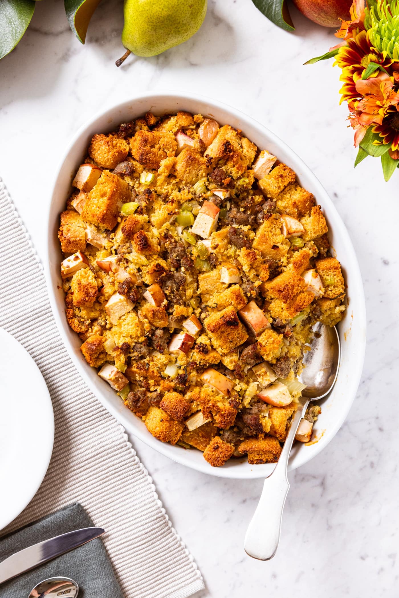 White marble surface with oval baking dish filled with cornbread stuffing with metal spoon holding a scoop of the stuffing before putting on a plate.