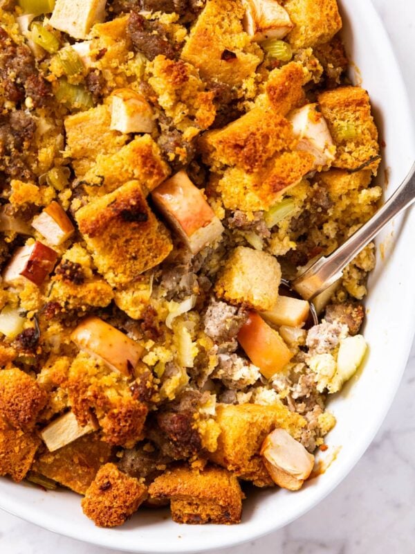 White baking dish filled with baked sausage cornbread stuffing with metal spoon resting inside with scoop of interior showing the apples and sausage also in the recipe.