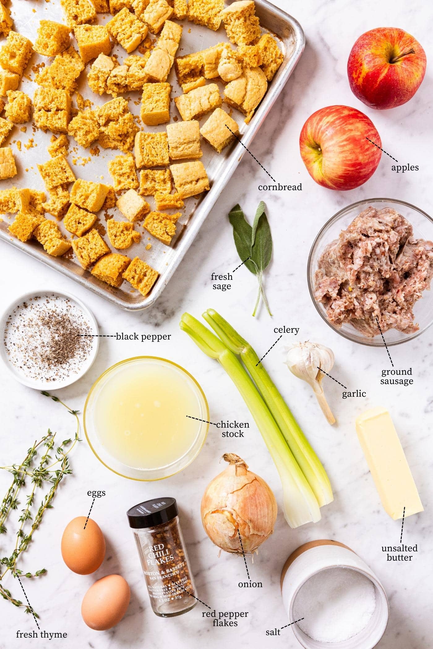 White marble surface filled with all of the ingredients needed to make cornbread stuffing including dried cornbread, apples, celery, onion, sausage, eggs, herbs, and more.