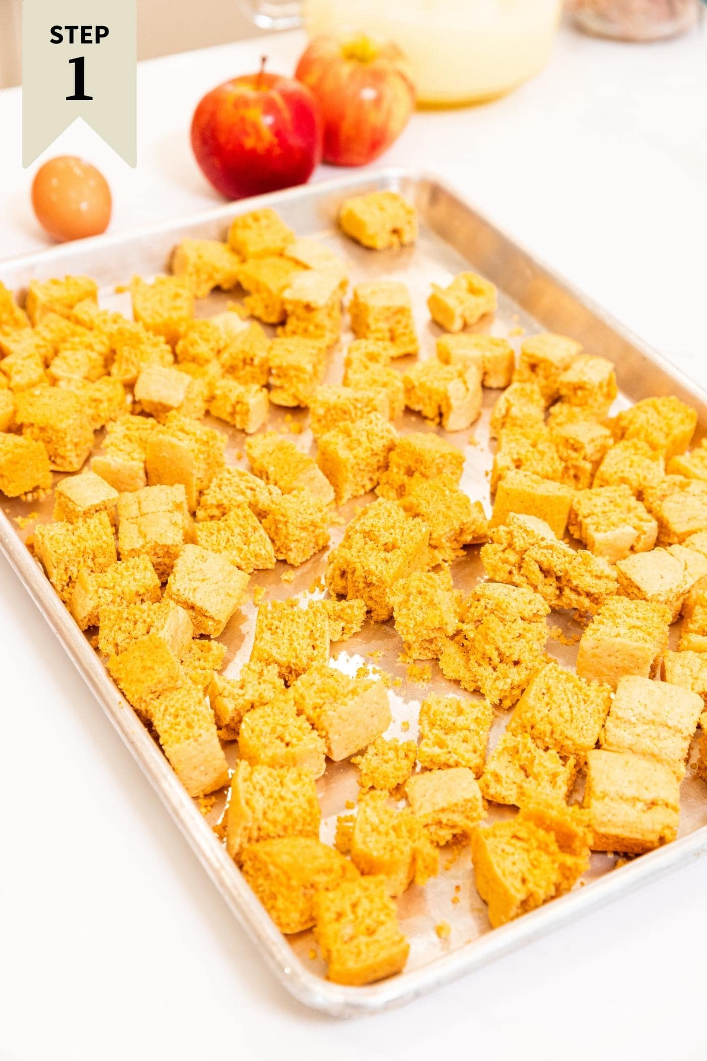 White countertop filled with a baking sheet with cubes of dried cornbread on it with apples and other stuffing ingredients in the background.