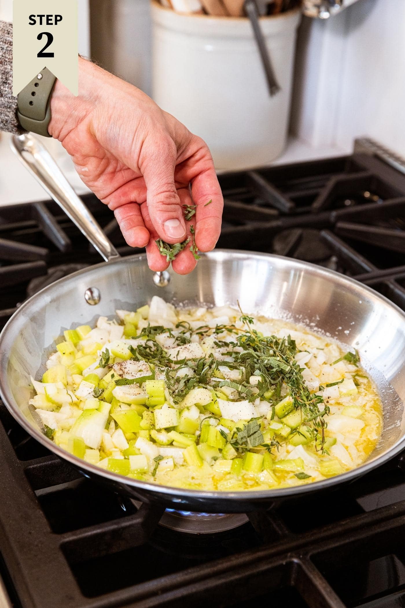 Metal skillet on stovetop filled with diced onion and celery with pieces of herbs being sprinkled on top while cooking in melted butter.