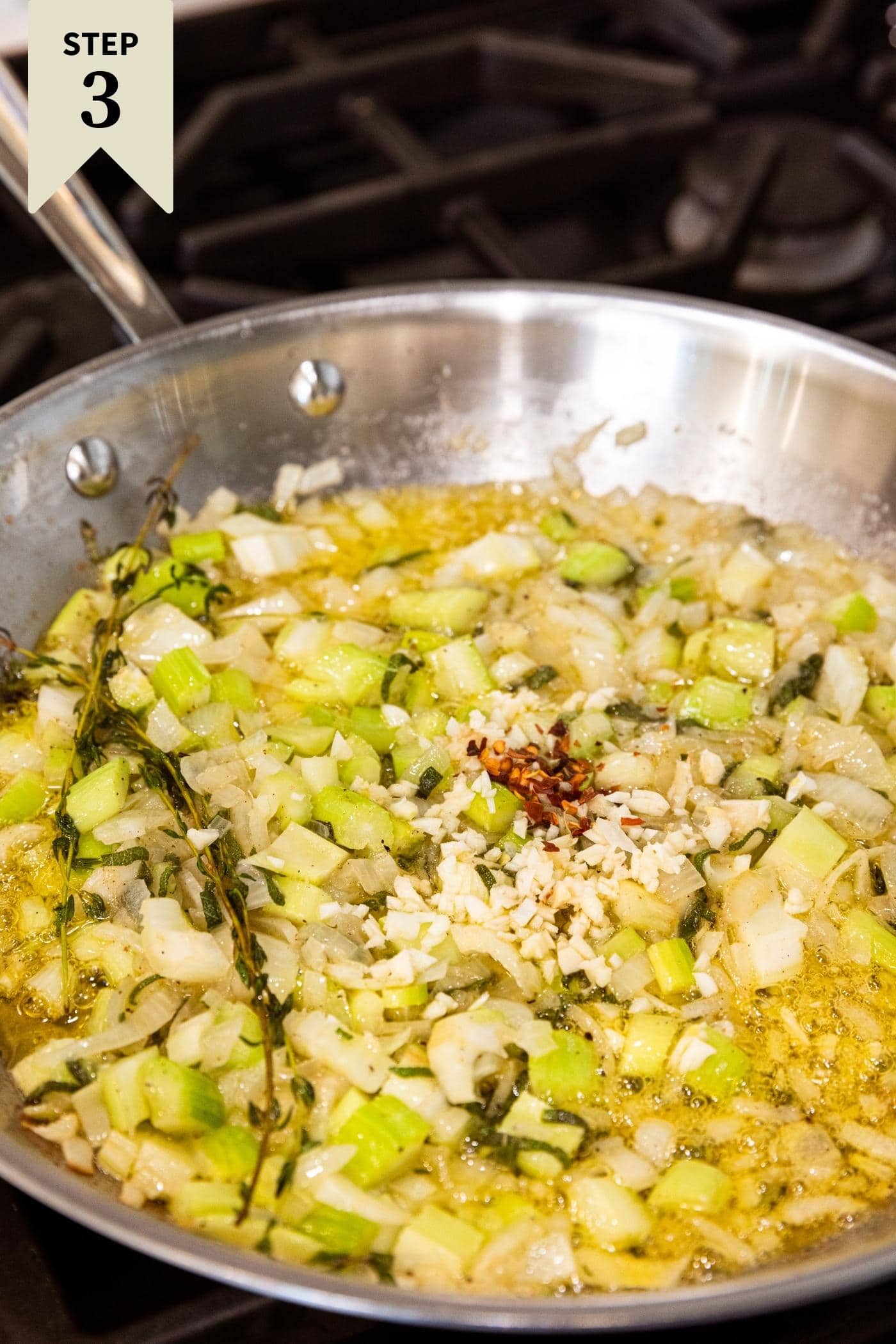 Metal skillet sitting on stovetop filled with diced onion and celery in melted butter with herbs and garlic and red pepper flakes sitting on top before being mixed together.