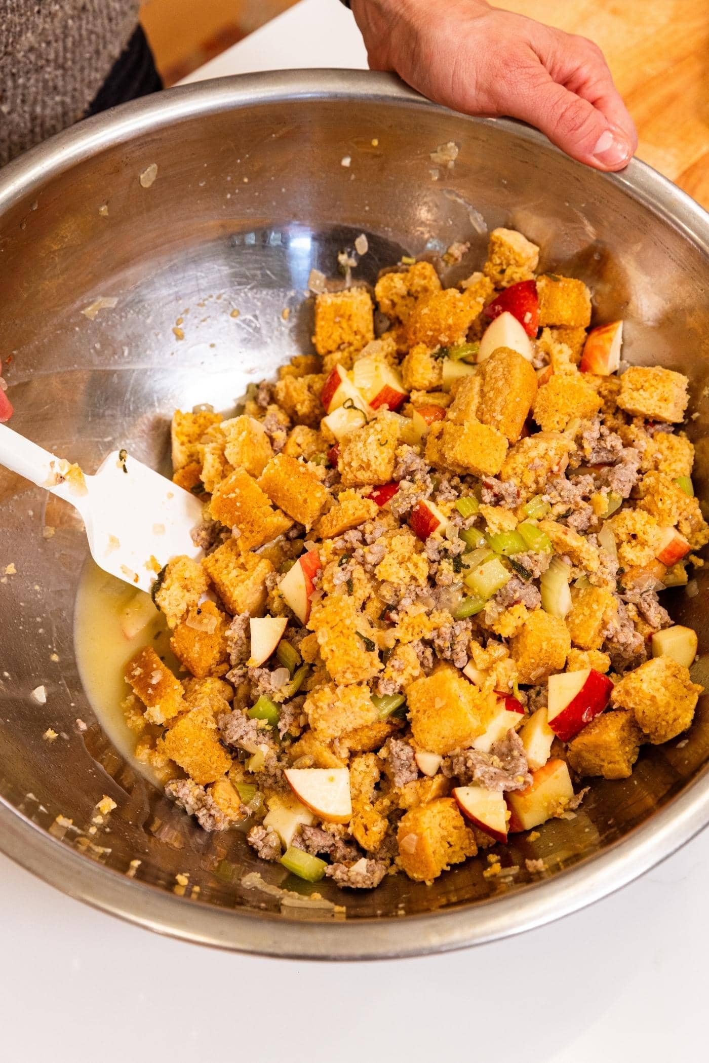 Hand stirring together cornbread with browned sausage, apple pieces, and chicken stock with white plastic spatula in large metal bowl.