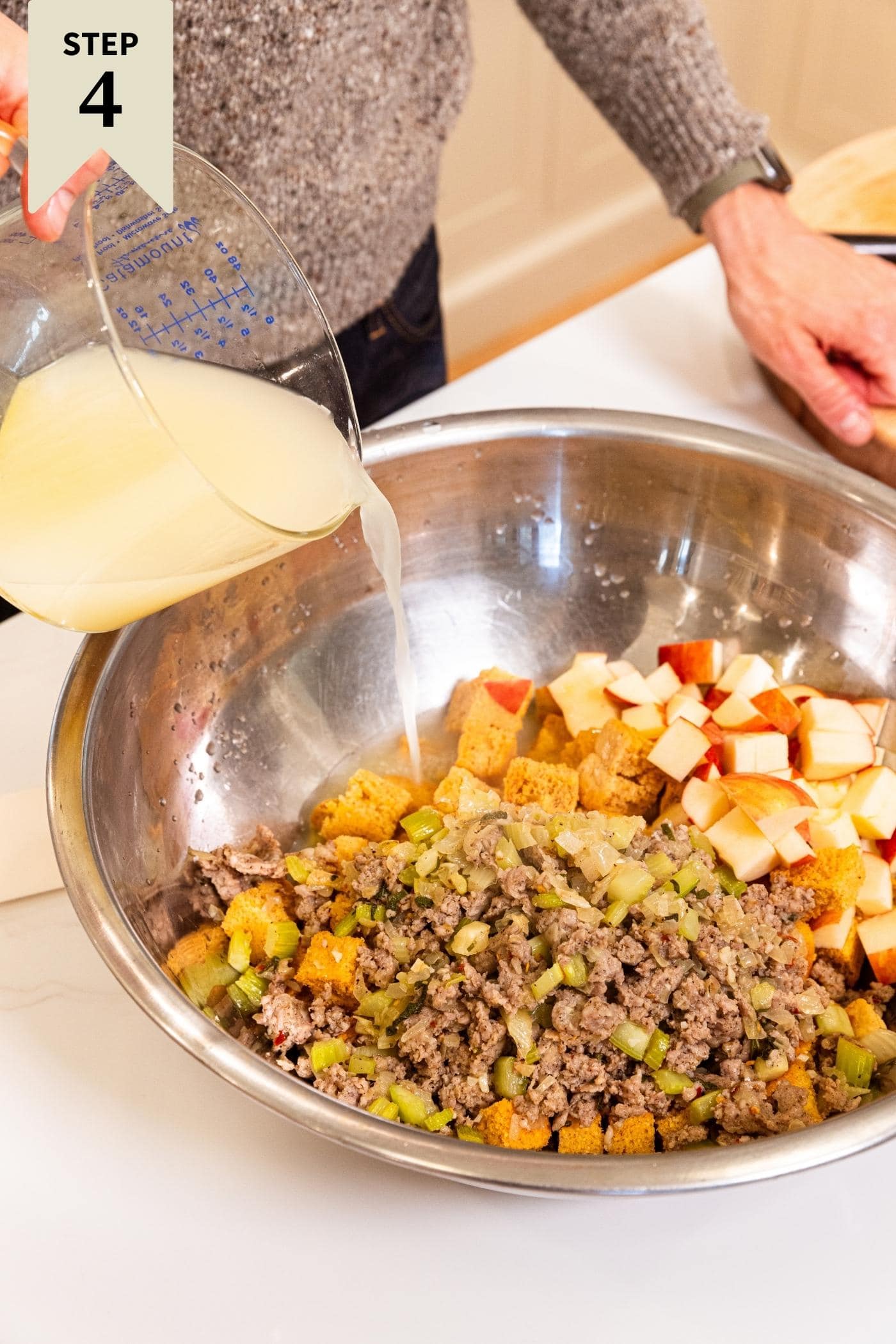 Large metal bowl filled with cornbread cut into cubes, chopped apple, and sausage mixture with hand pouring in light yellow colored chicken stock over the entire mixture.