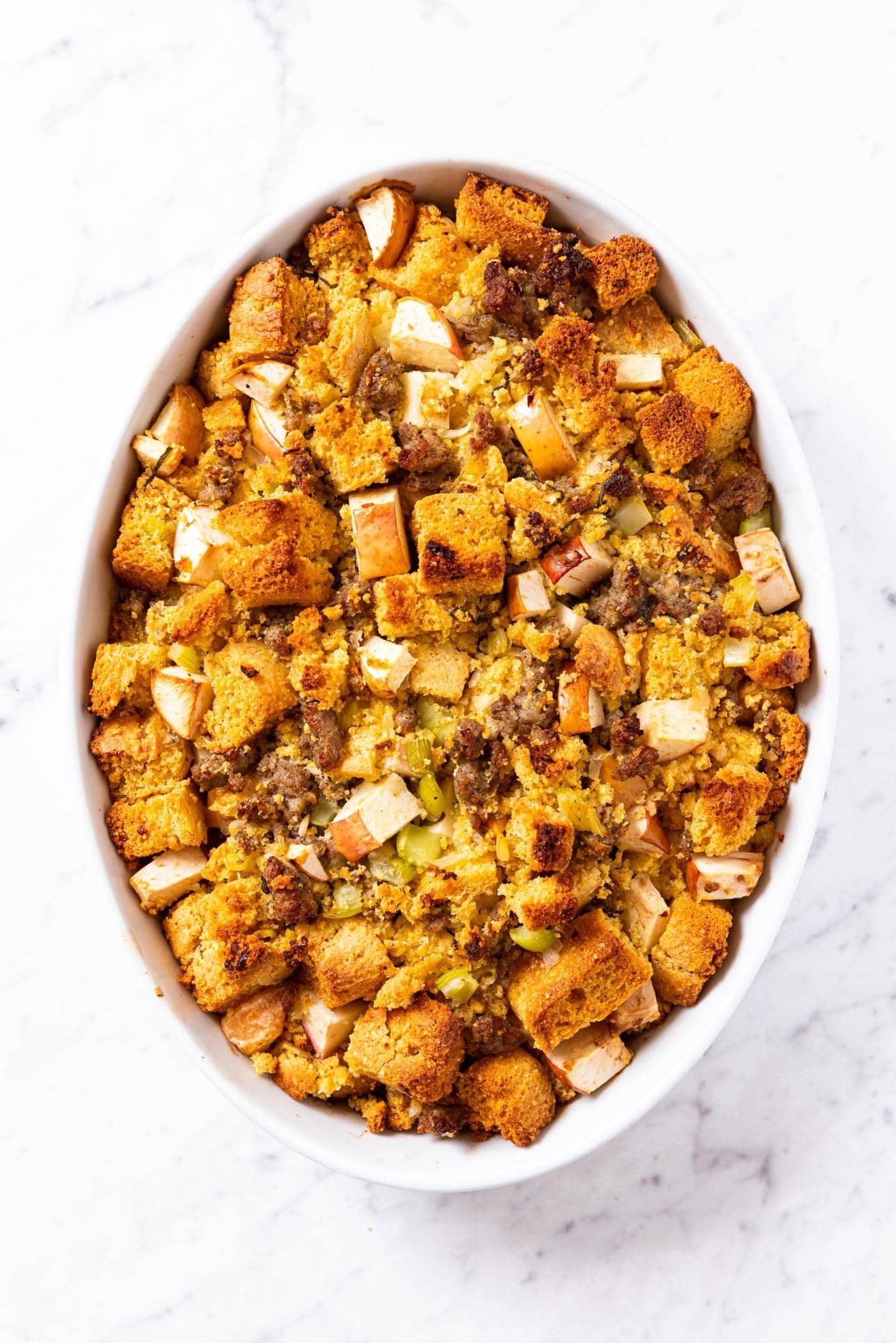 Apple and sausage cornbread stuffing mixture sitting on marble countertop after coming out the oven after baking with browned top showing crispy texture.