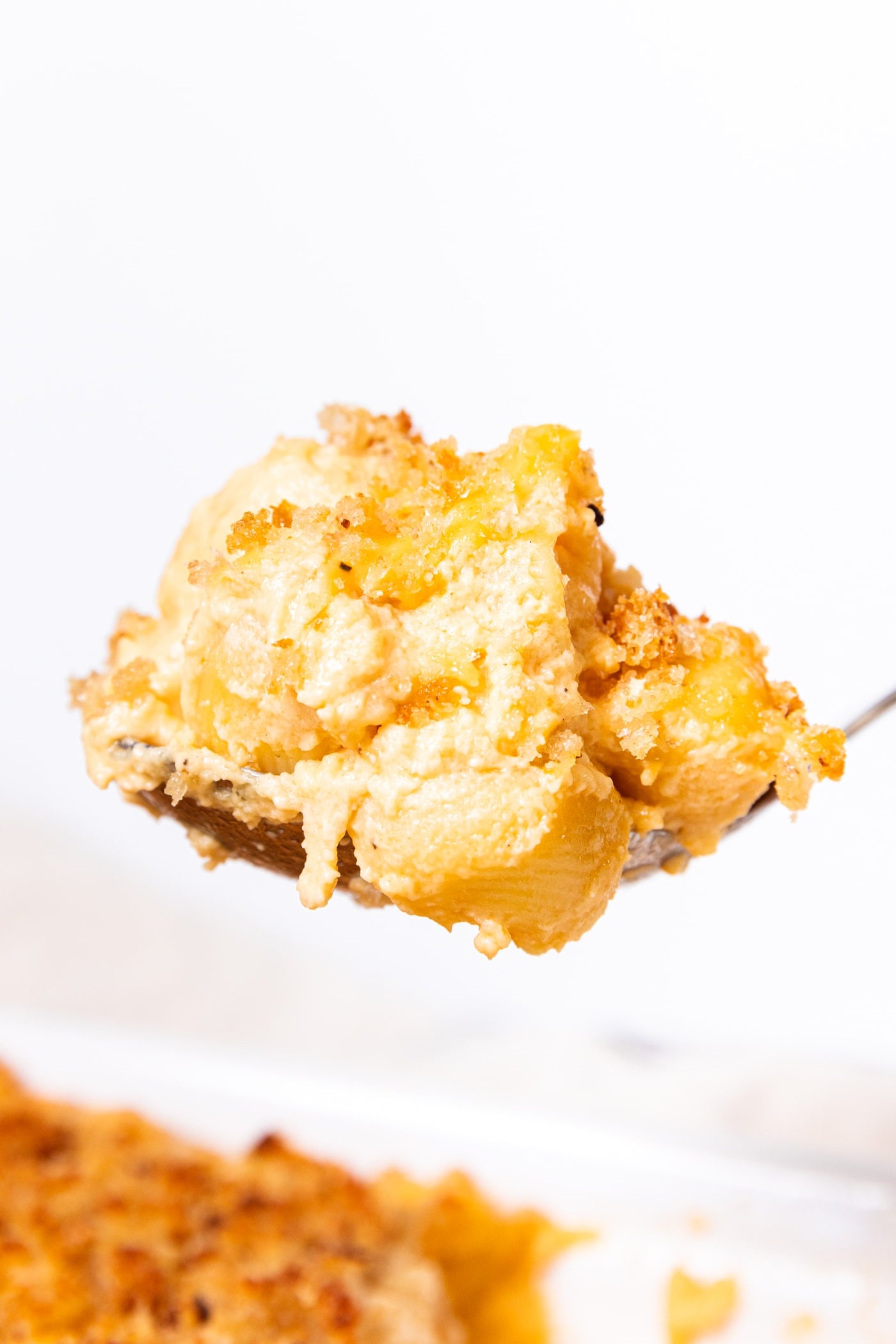 Spoon holding serving of baked macaroni & cheese showing the elbow macaroni and cheese mixed together with rest of casserole below in white pan.