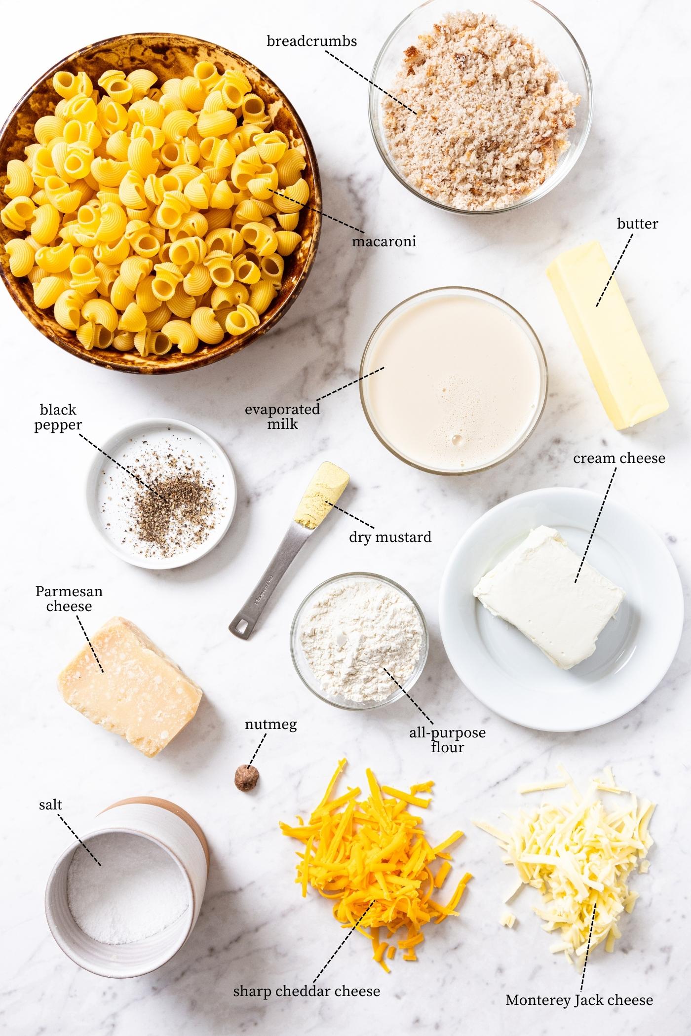 Marble surface filled with all ingredients needed to make baked macaroni & cheese including pasta, cheeses, evaporated milk, breadcrumbs, spices, and more.