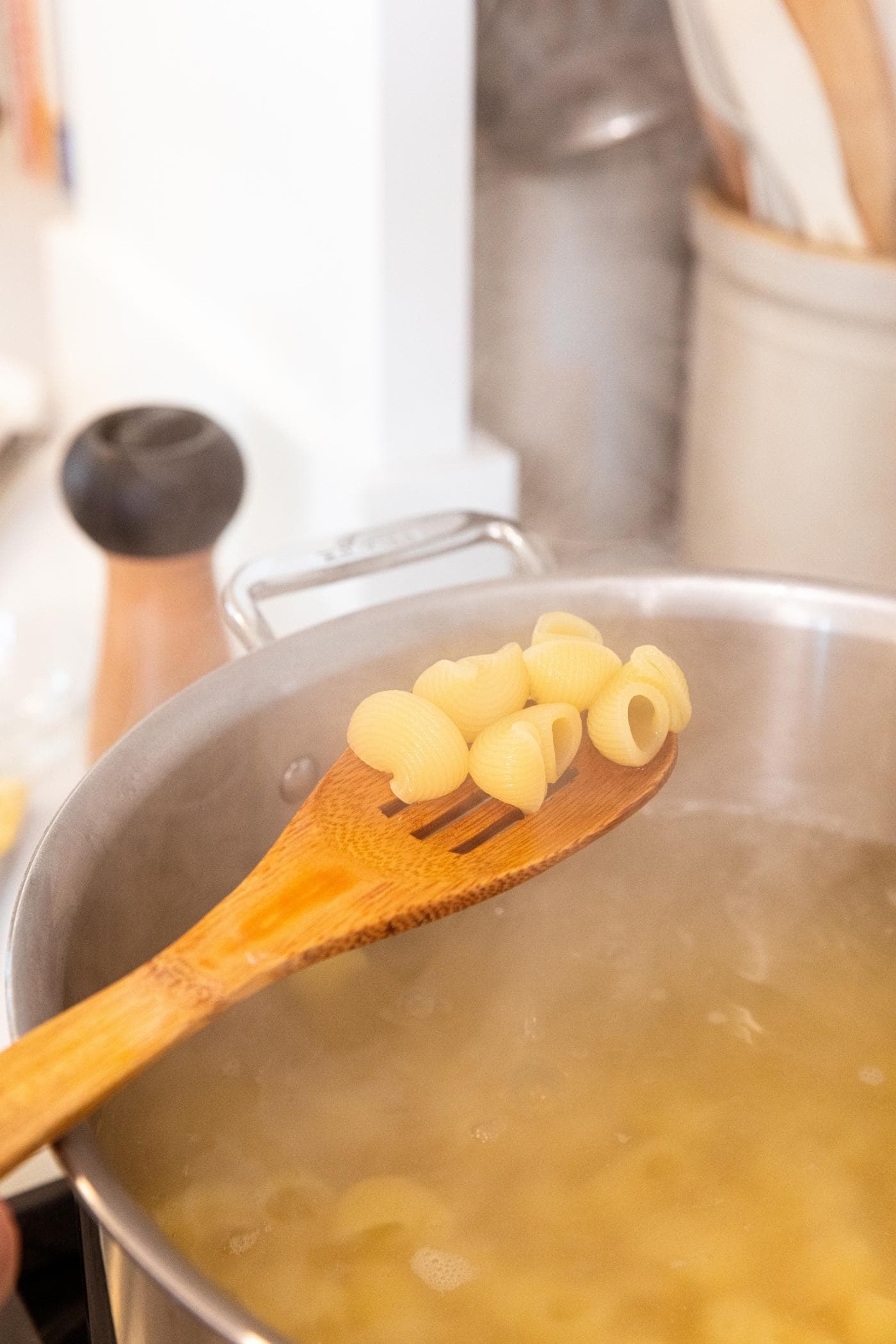 Wooden spoon holding cooked macaroni above surface of water with more macaroni in water below in stockpot on stove.