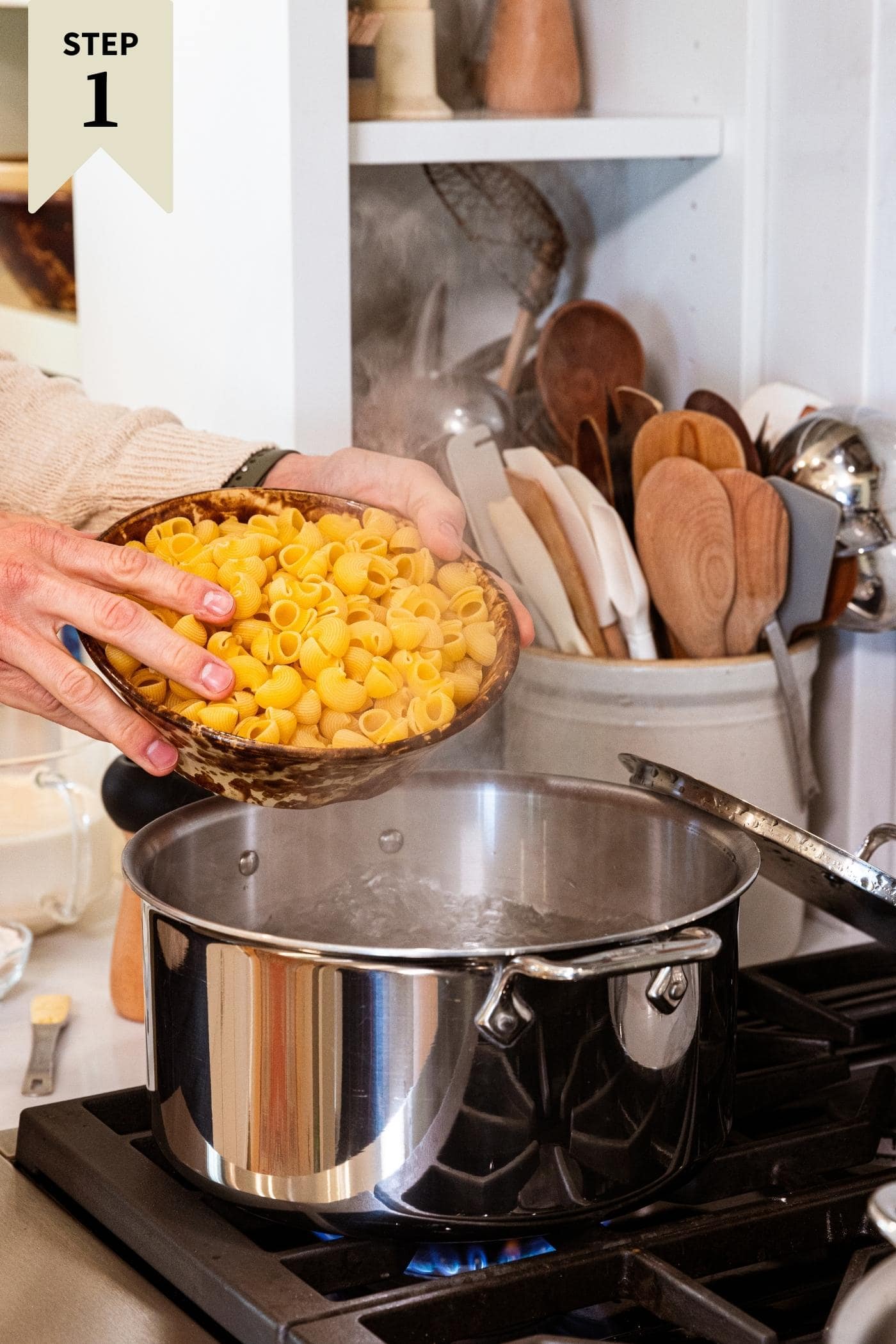 Two hands holding brown bowl about to pour elbow macaroni into pot filled with bowling water on stovetop.