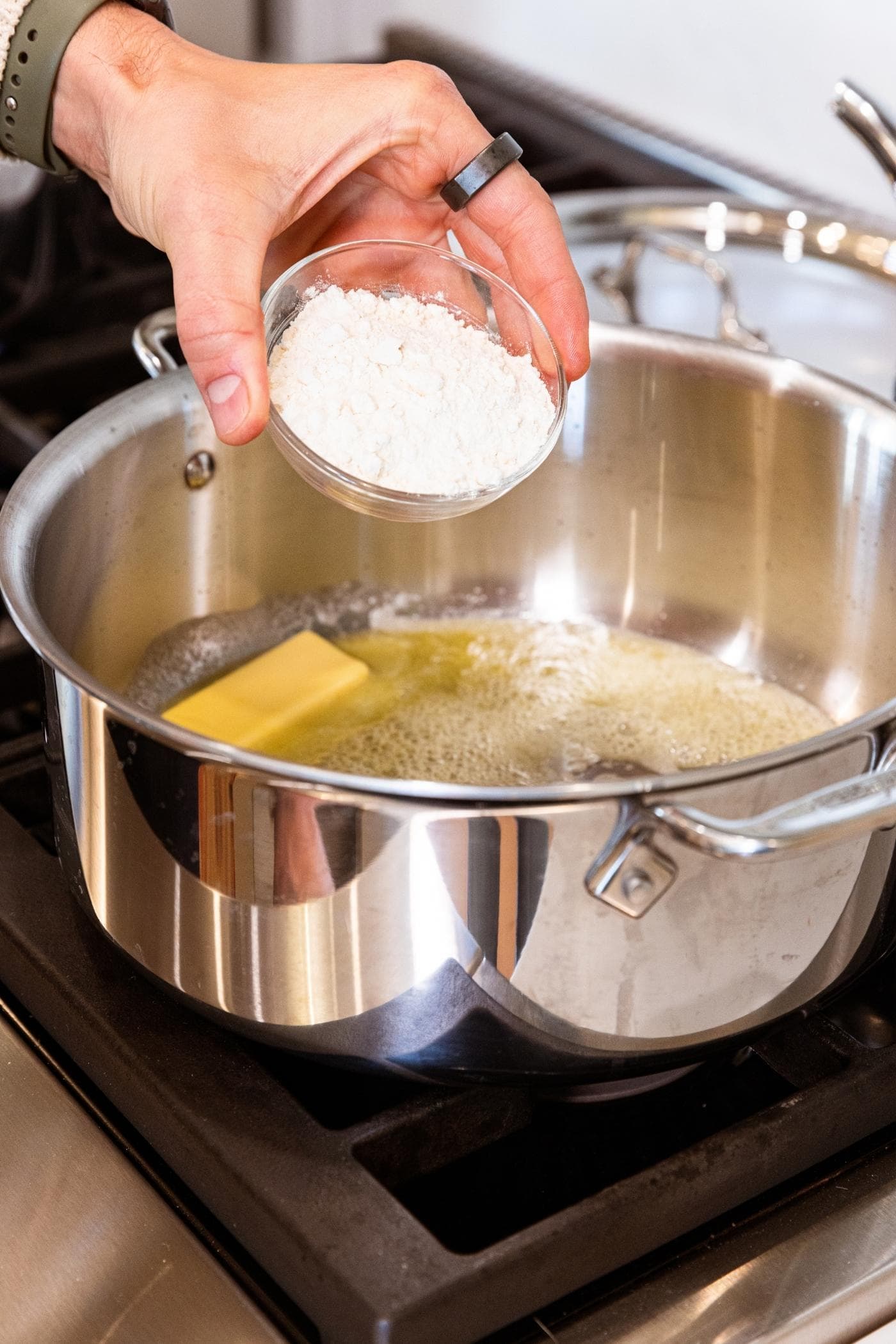 Hand holding small glass bowl filled with flour about to pour into stockpot on stovetop already filled with melting butter.