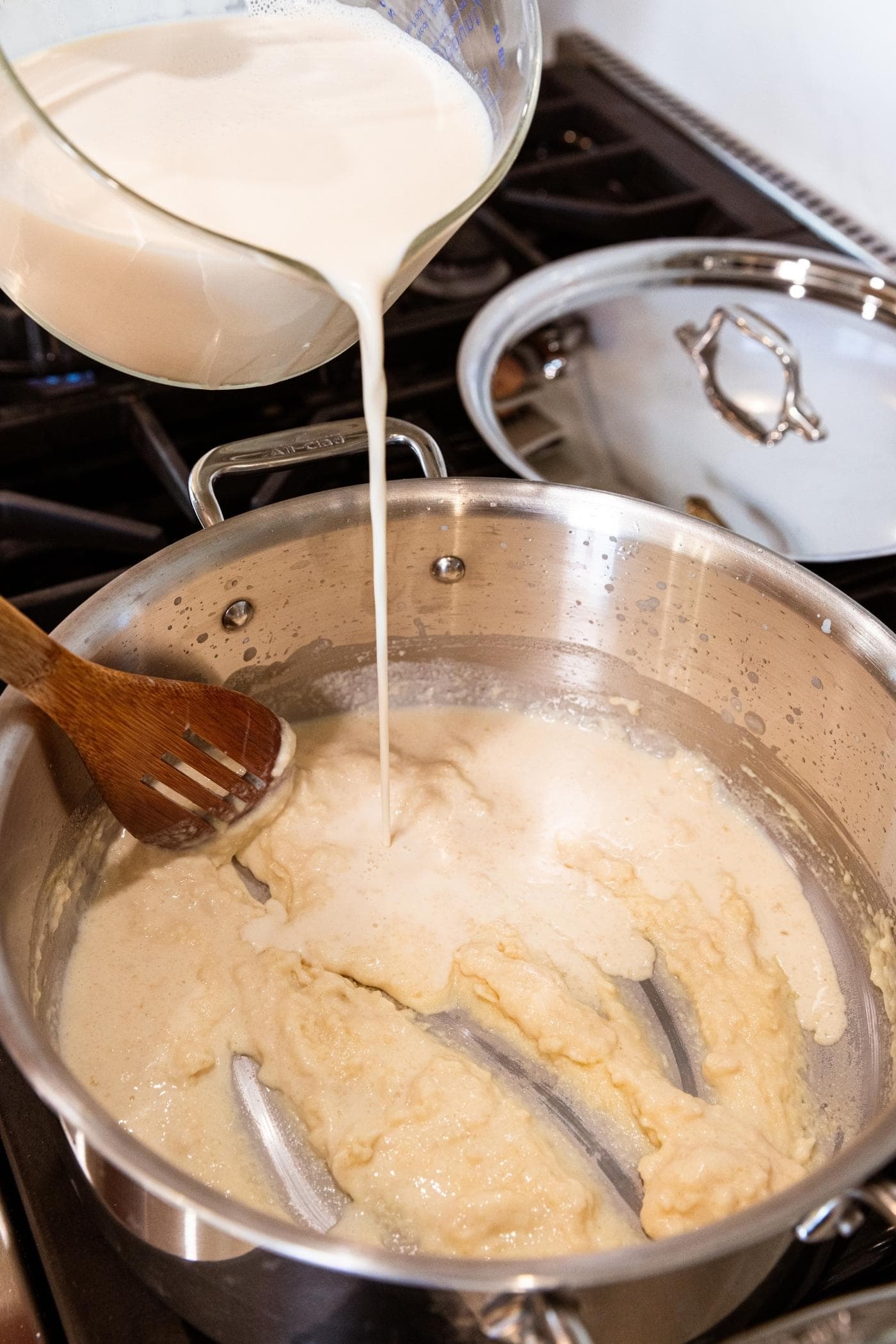 Large glass measuring cup pouring evaporated milk into roux mixture in large stockpot on stovetop with wooden spoon also in pot about to stir.