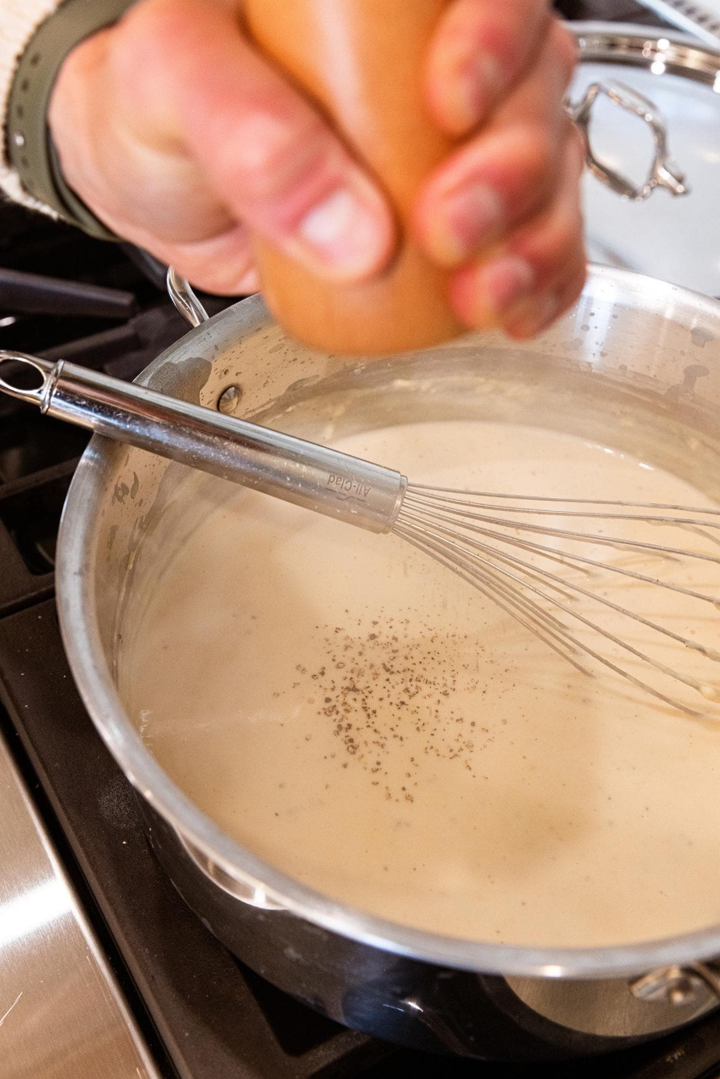 Hand grinding black pepper from brown colored pepper grinder on top of milk mixture with whisk resting on side of metal stockpot.
