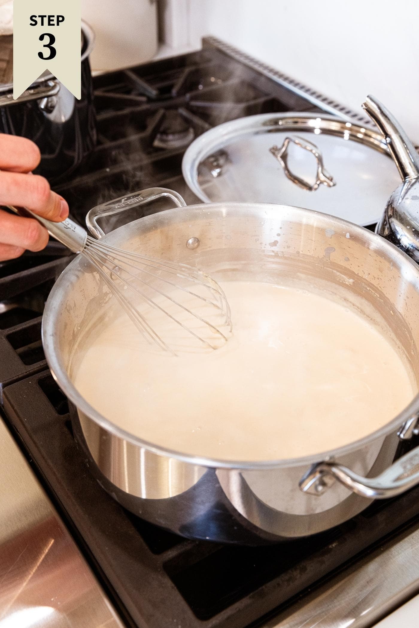 Hand holding whisk stirring together milk mixture in large stockpot over heat on stovetop with steam coming off the top.