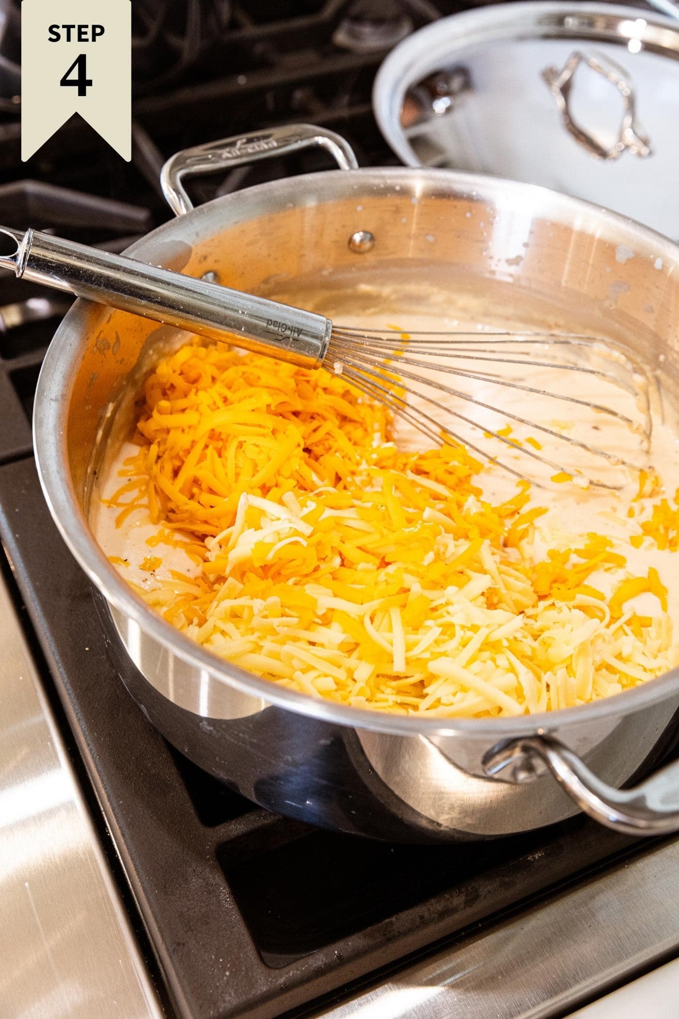 Shredded cheese sitting on surface of milk mixture in large stockpot on stovetop with whisk resting against the side of the pot.