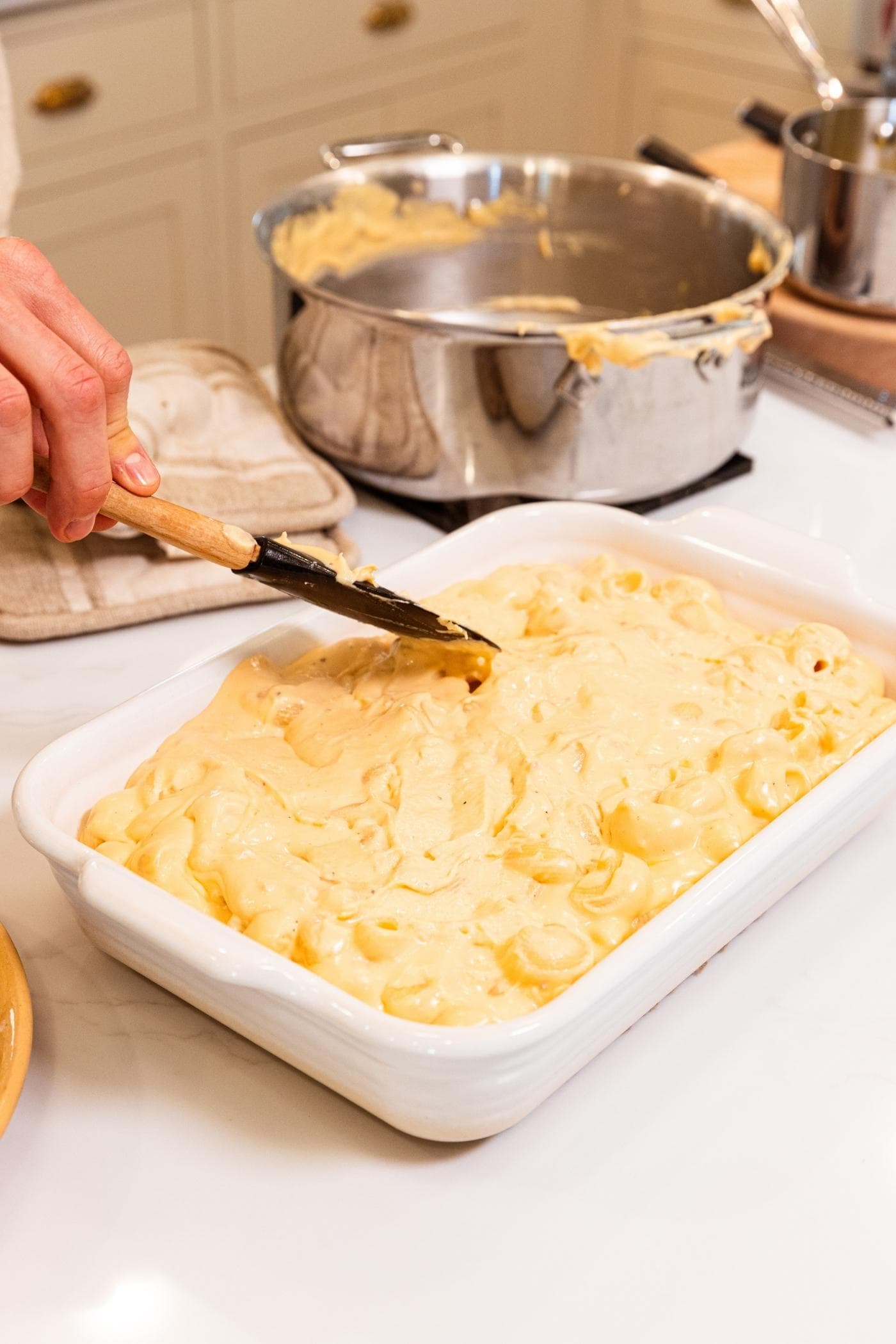 Hand using spatula to smooth out top of macaroni & cheese mixture in white baking dish on white countertop.