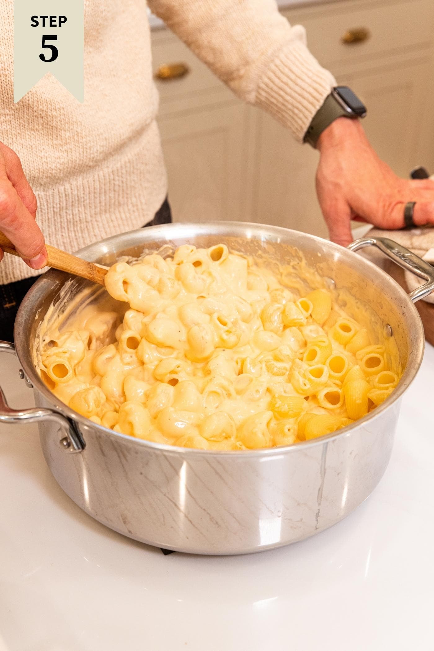 Hand stirring together cheese with elbow macaroni in large metal stockpot on white countertop with creamy colored cabinets in background.