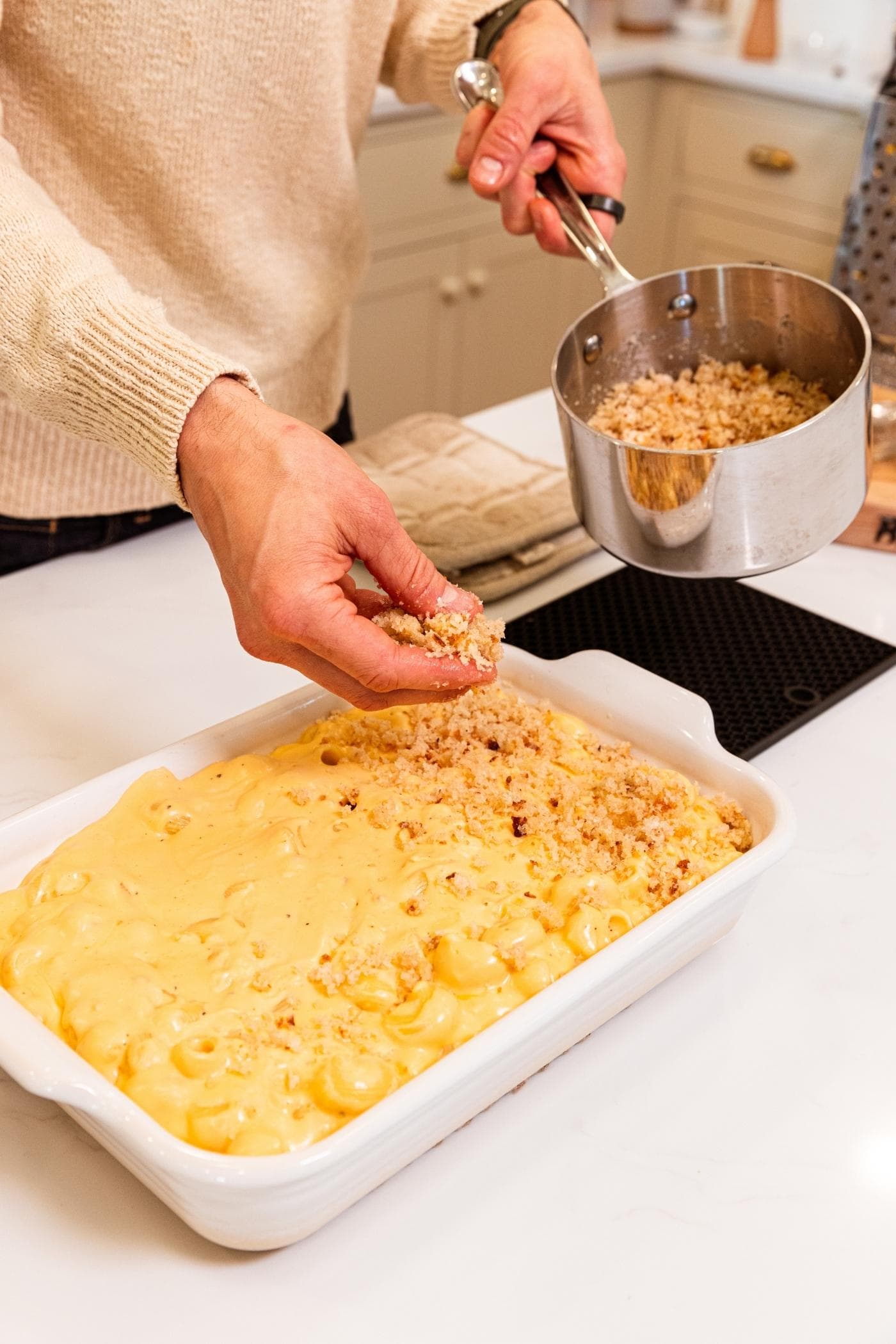 Hand sprinkling breadcrumb mixture on top of macaroni & cheese mixture in white baking dish from small metal kettle with more breadcrumb mixture.