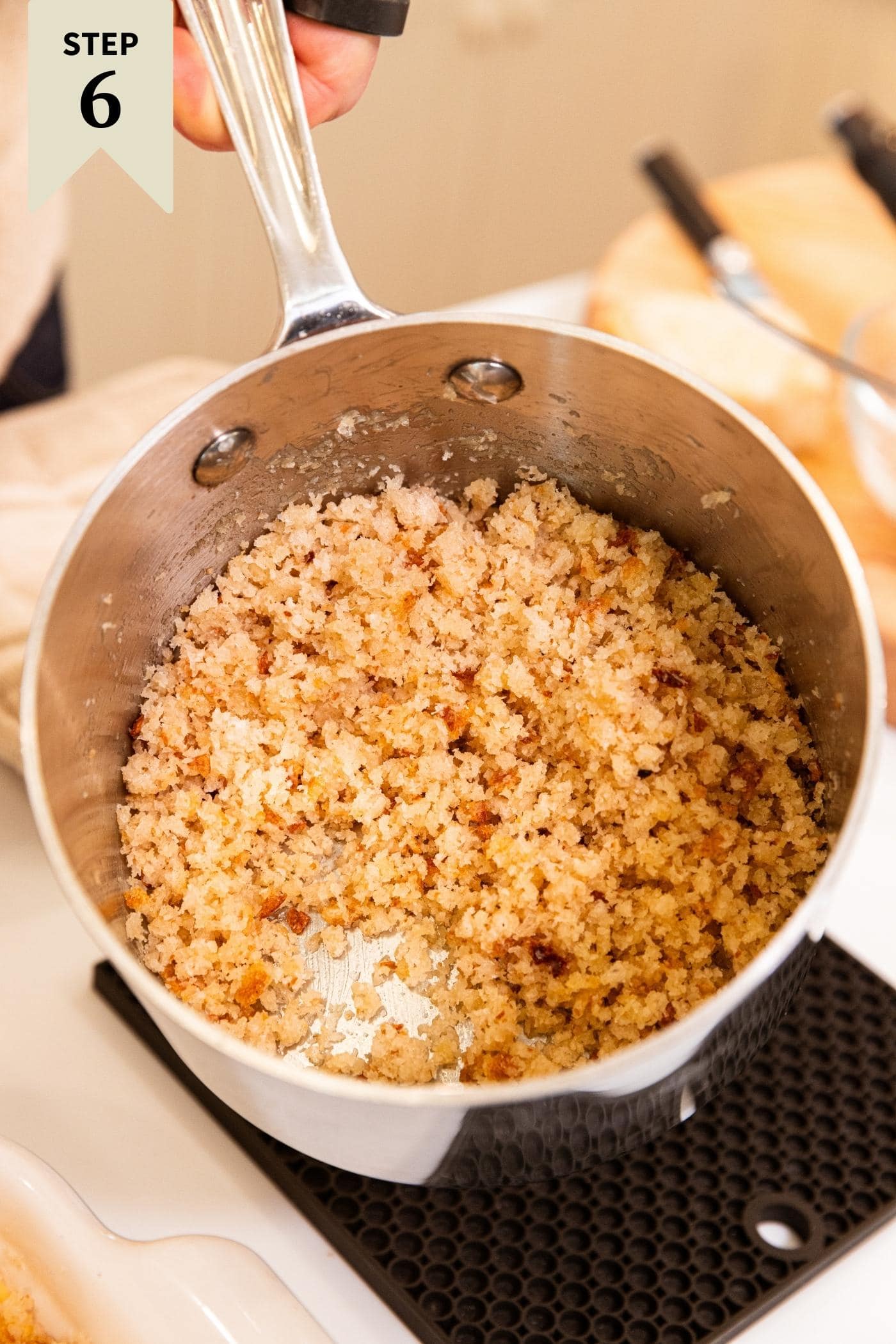 Hand holding metal kettle filled with breadcrumbs mixed with breadcrumbs all mixed together to form breadcrumb topping.