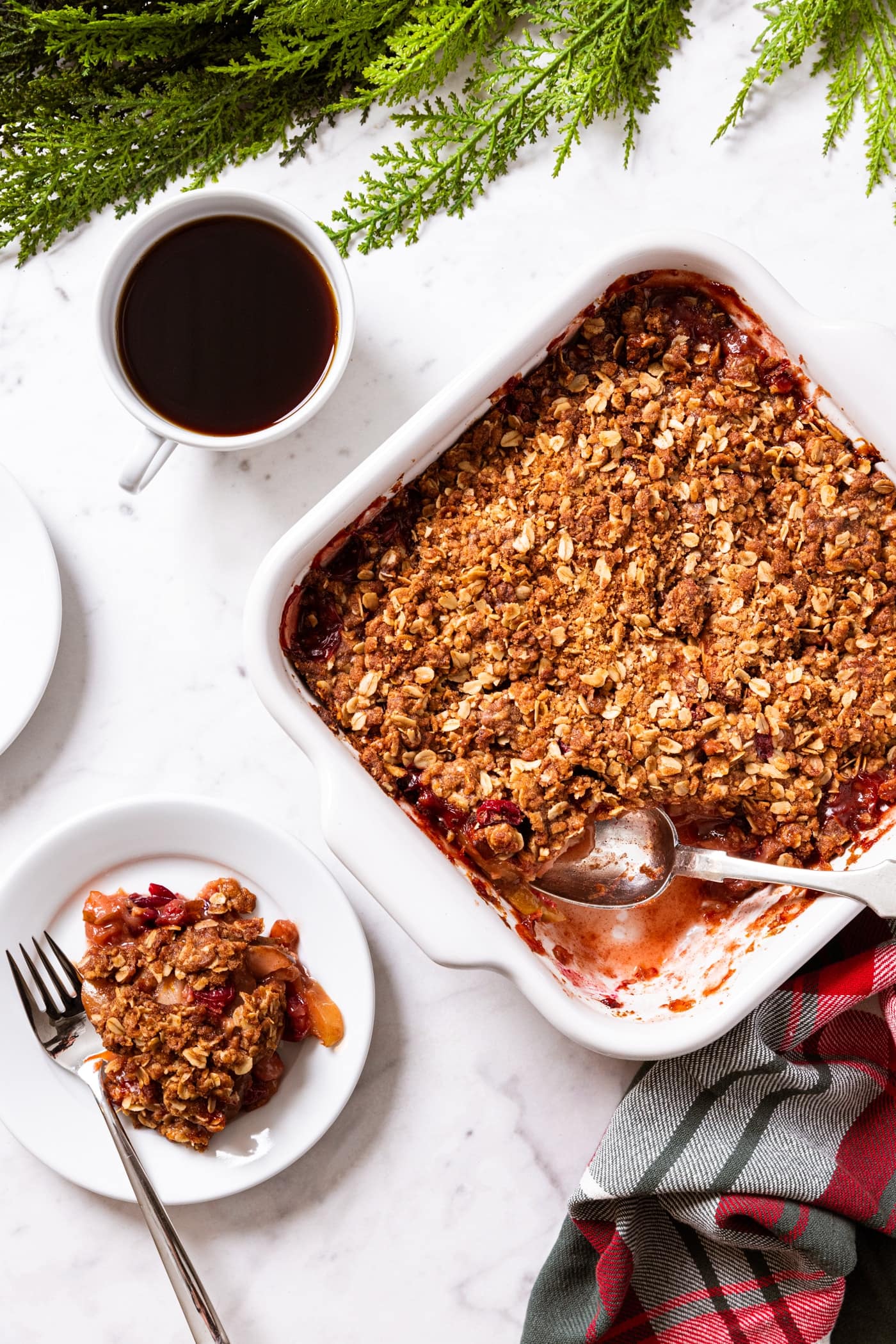 White marble surface with white baking dish filled with baked cranberry crisp with cup of coffee and white plate filled with a scoop of the crisp.