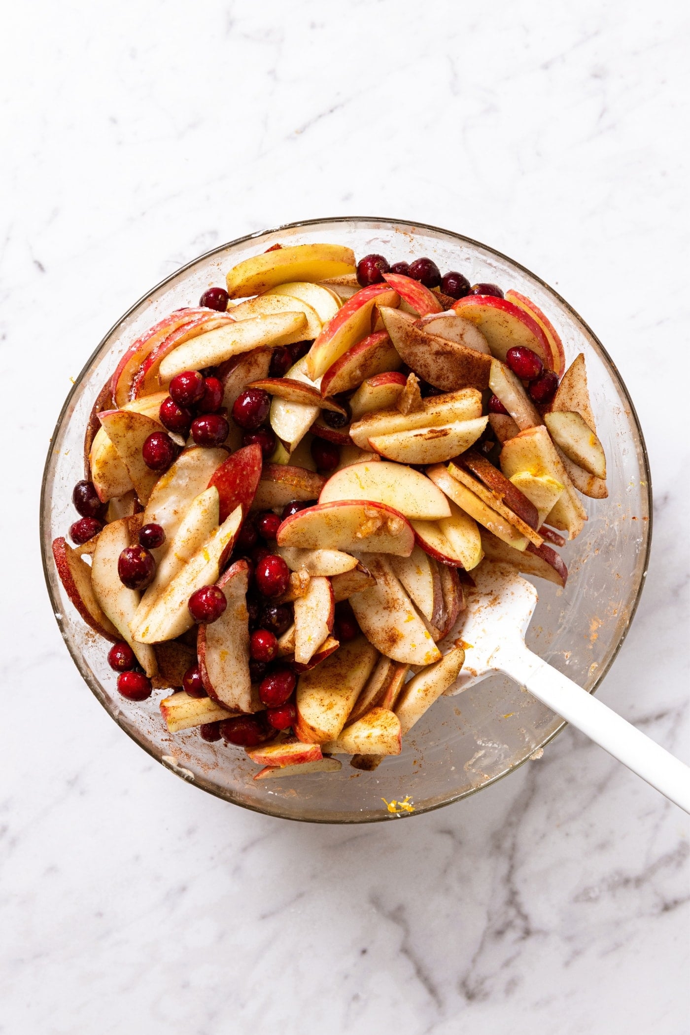 White marble surface with glass bowl full of mixed ingredients including apples, pears, and cranberries to make a cranberry crisp with plastic spatula.