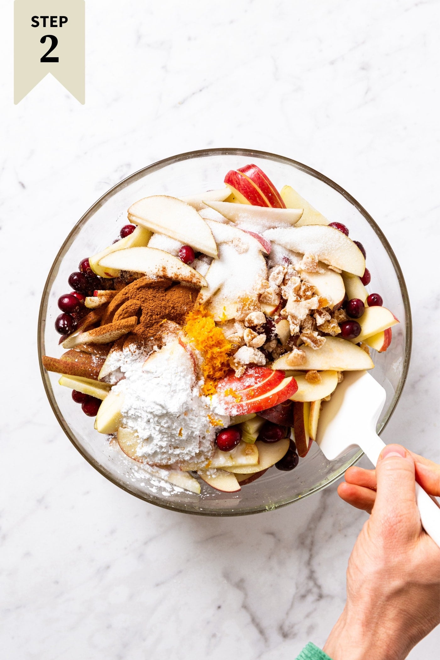 White marble surface with glass bowl filled with all ingredients needed to make a cranberry crisp with hand using white plastic spatula to mix everything.