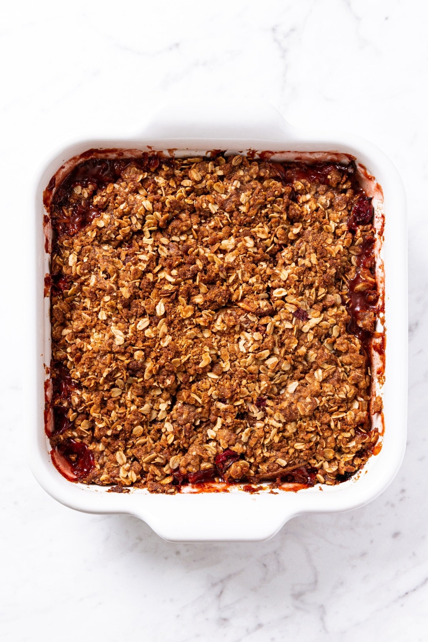 Cranberry crisp in white baking dish sitting on marble surface after being baked in the oven until the top is crispy and browned.
