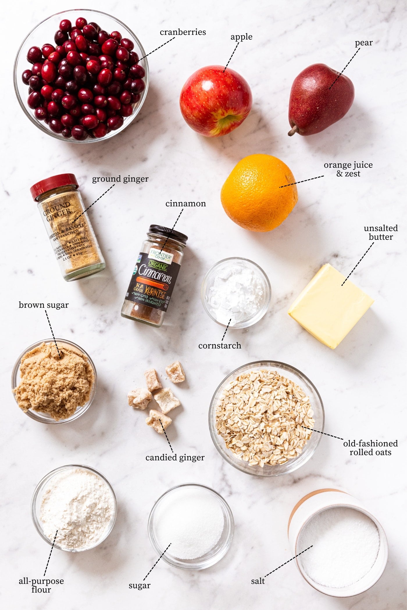 White marble surface containing all ingredients needed to make a cranberry crisp including cranberries, pear, apple, sugar, butter, orange, spices, and more.