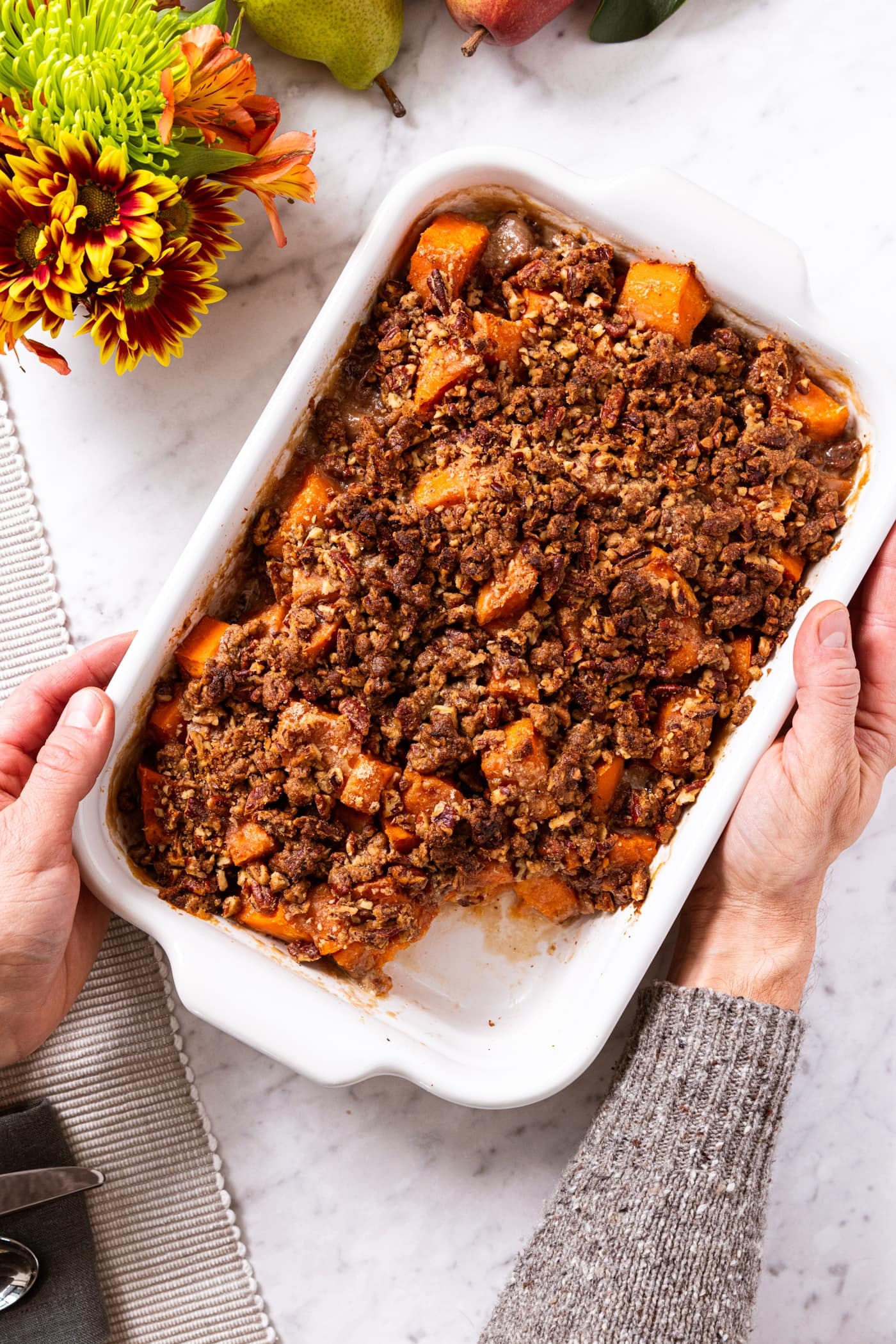 Two hands setting white baking dish filled with sweet potato casserole on white marble surface with flowers and magnolia garland.