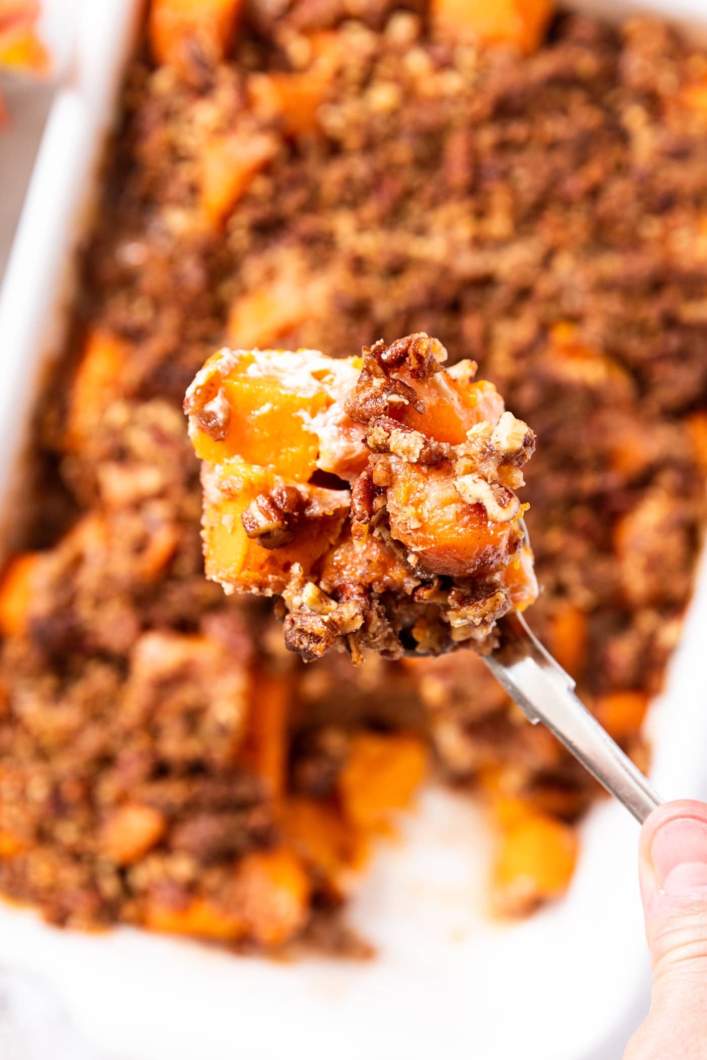 Spoon holding scoop of sweet potato casserole with chunks of orange colored sweet potatoes topped with pecan streusel topping with rest of baking dish sitting below.