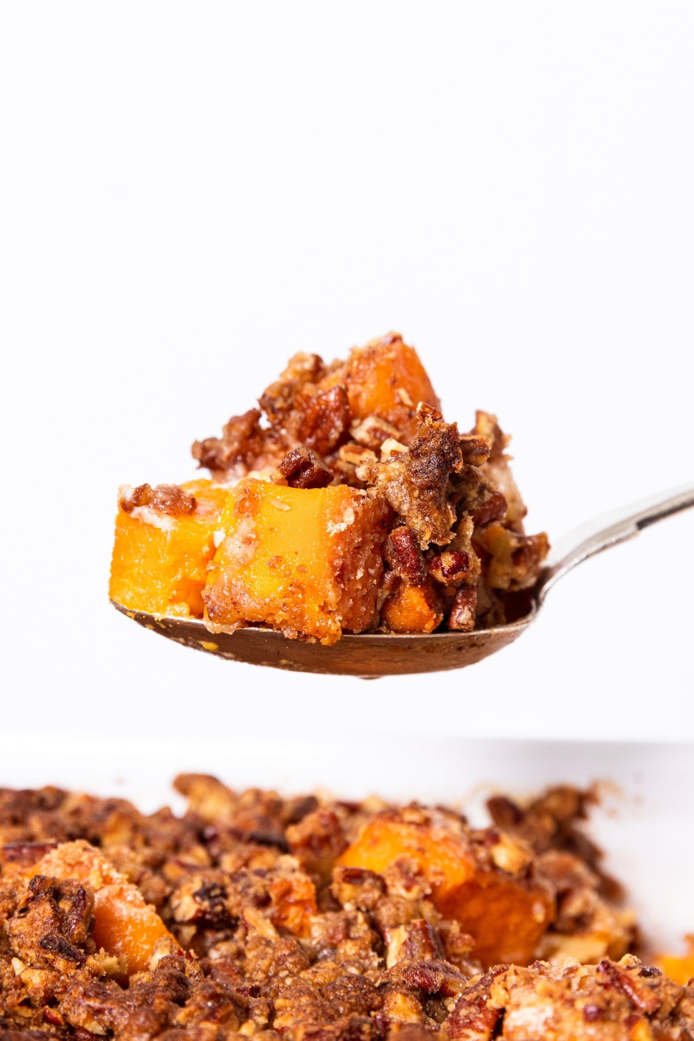Spoon holding scoop of sweet potato casserole with pecan streusel topping on top with rest of casserole in white baking dish below.