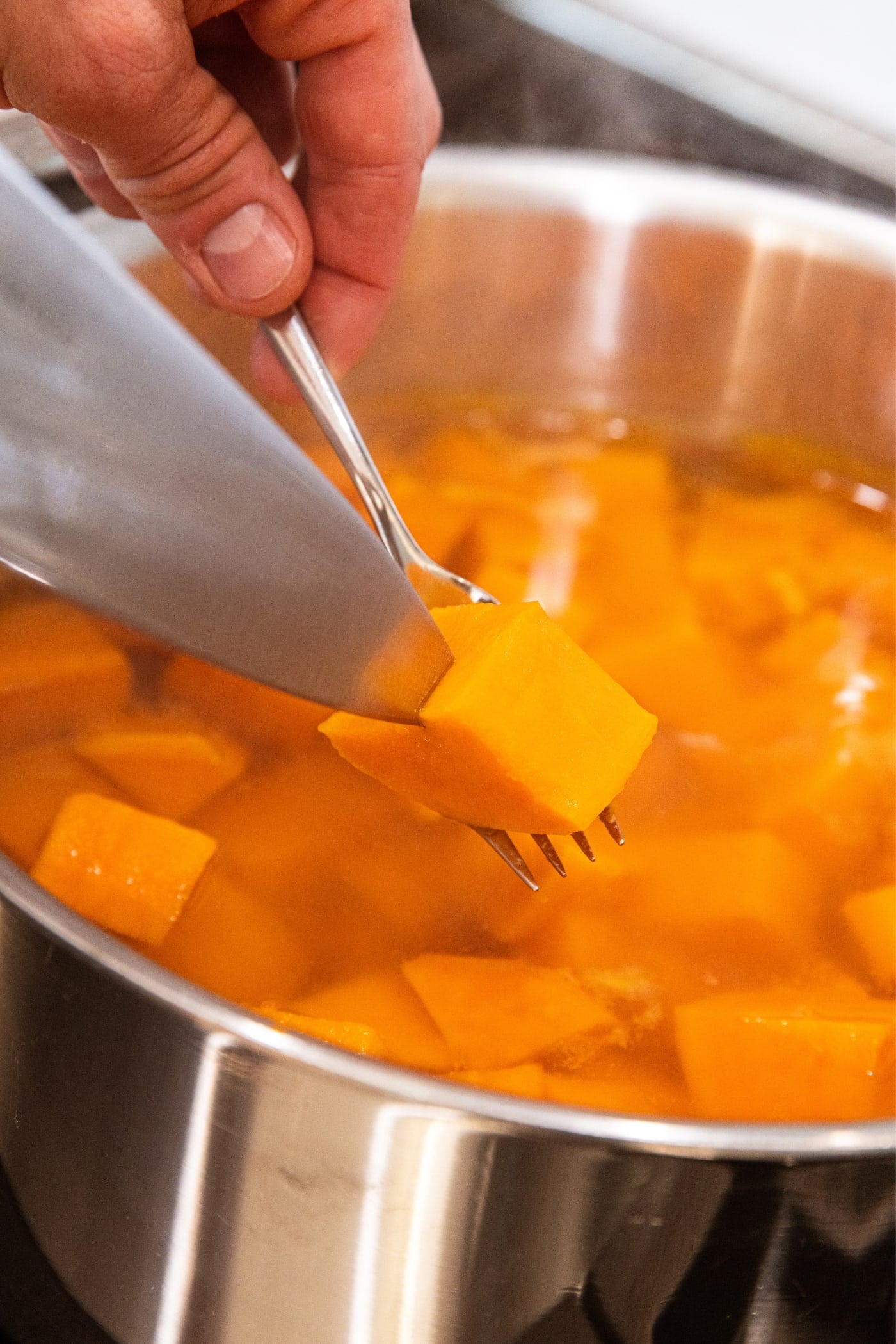 Hand holding knife inserting it into the center of a sweet potato being held on a fork over a kettle filled with water and more cooked sweet potatoes.