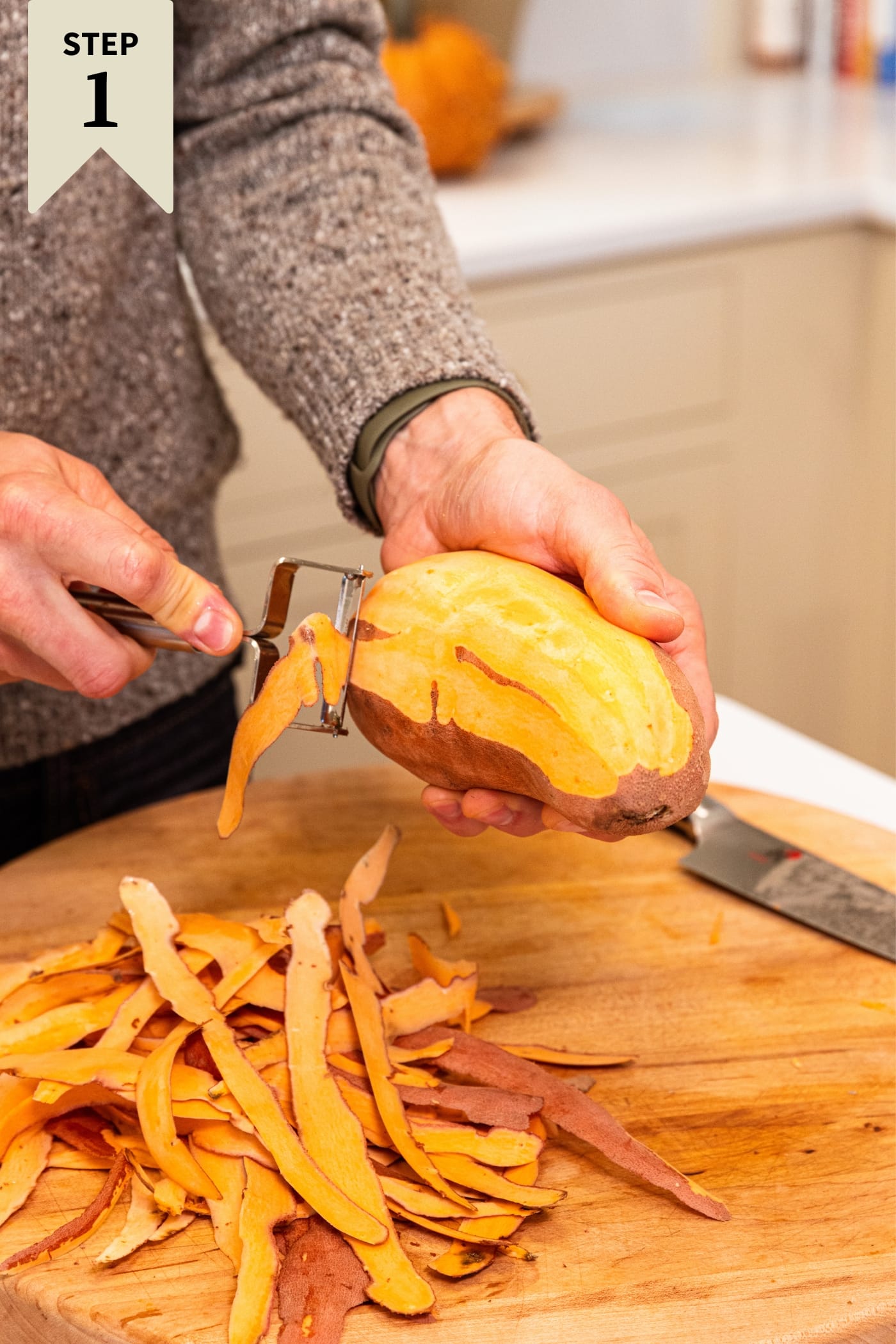 Hand using a vegetable peeler to peel an orange colored sweet potato over wood cutting board with knife and more peel all around.