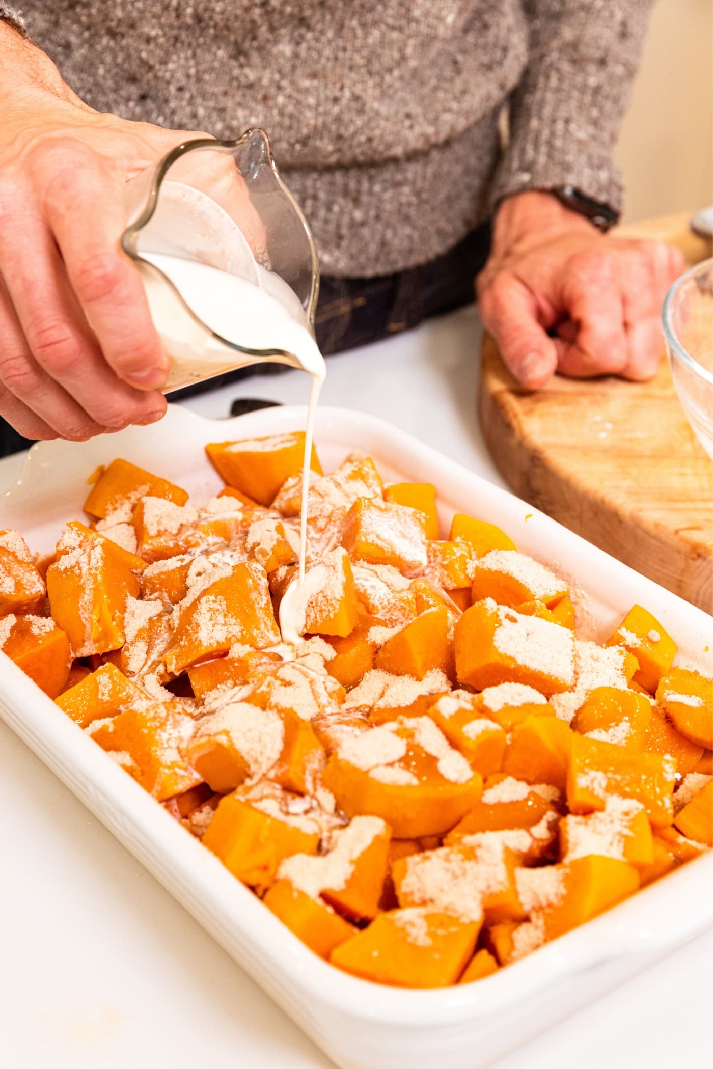 Hand holding glass container filled with heavy cream pouring into a baking dish filled with chunked sweet potatoes with white flour on top.