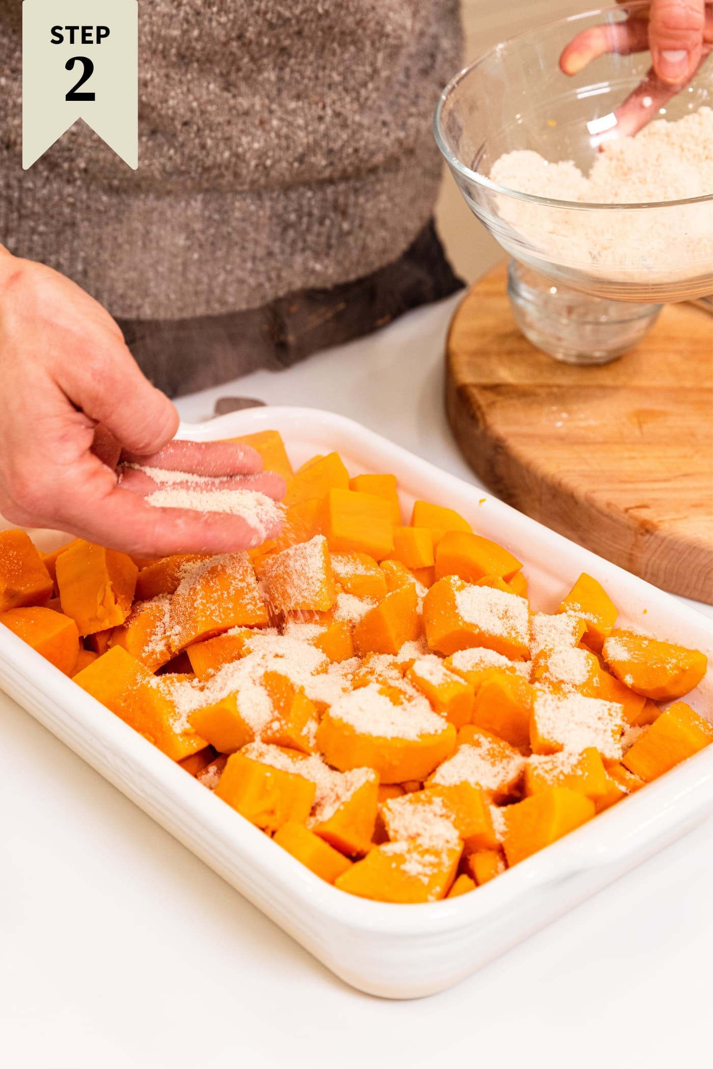 Hand sprinkling white flour mixture all over top of chopped sweet potatoes in white baking dish on white countertop with glass bowl filled with the rest of the flour mixture.