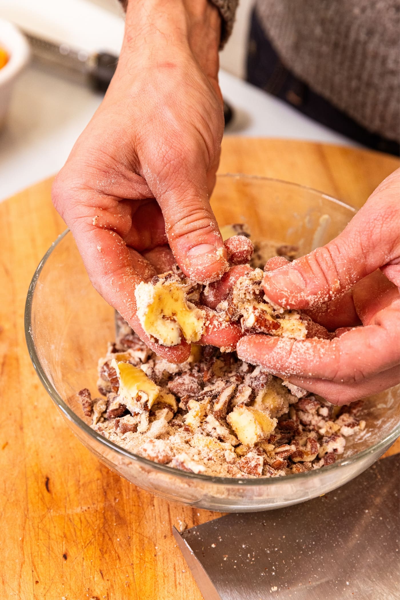 Hands holding pieces of butter squishing between finders over bowl filled with streusel topping mixture all on wood cutting board.