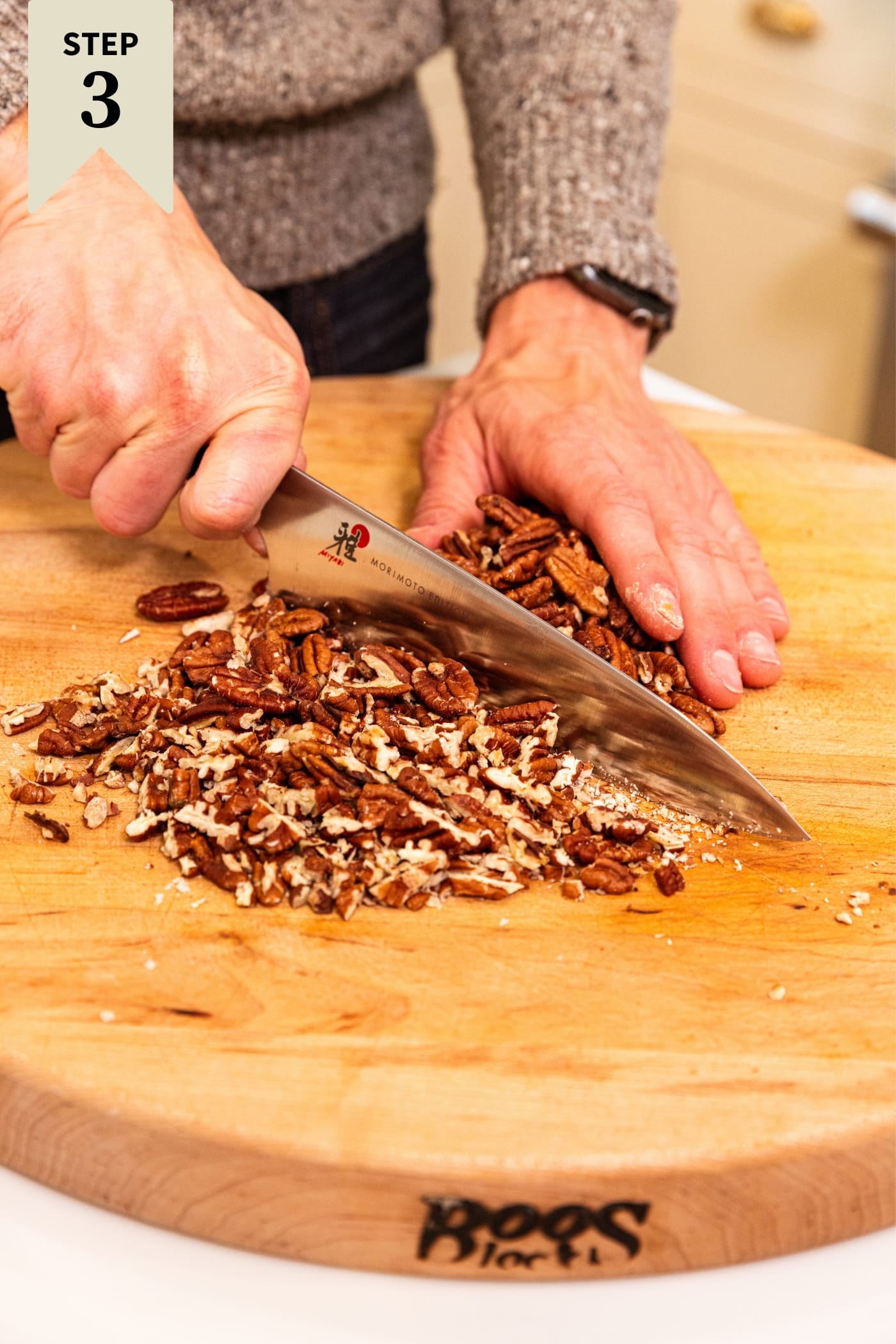 Hand holding knife chopping pecans on wood cutting board into small sized pieces.