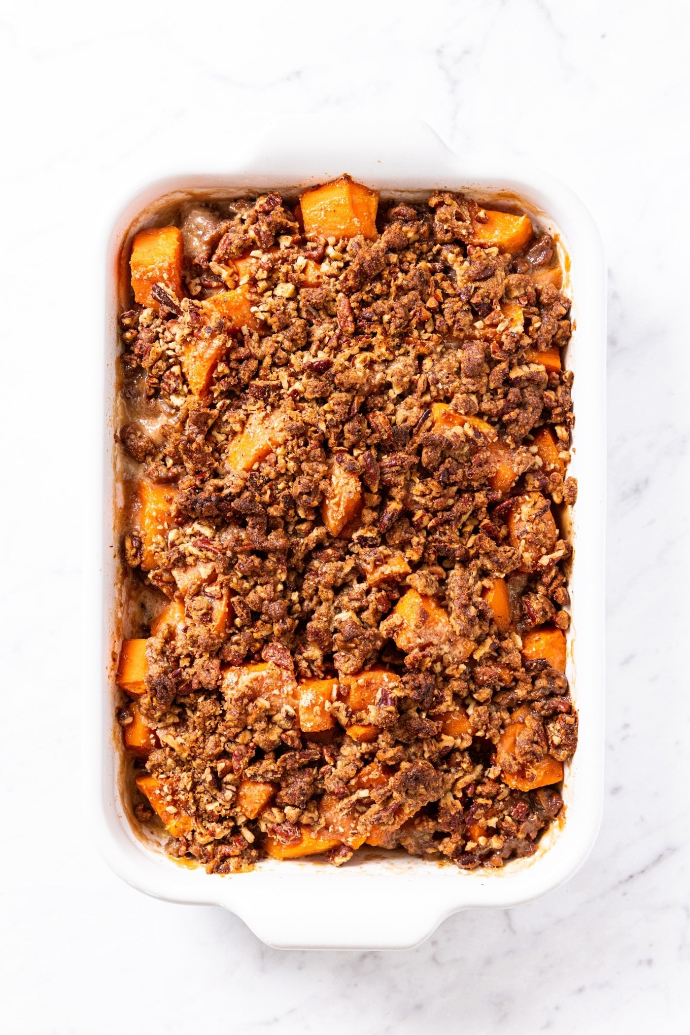 White marble surface with white baking dish sitting after coming out of the oven with browned texture on top with chunks of sweet potato peeking through.