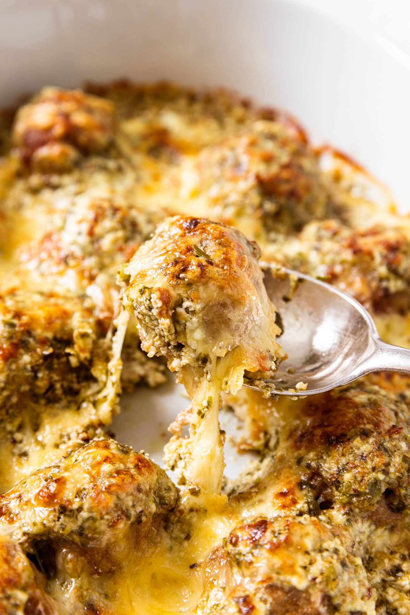 Spoon scooping up a meatball from the rest of meatballs in white baking dish with cheese pulling.