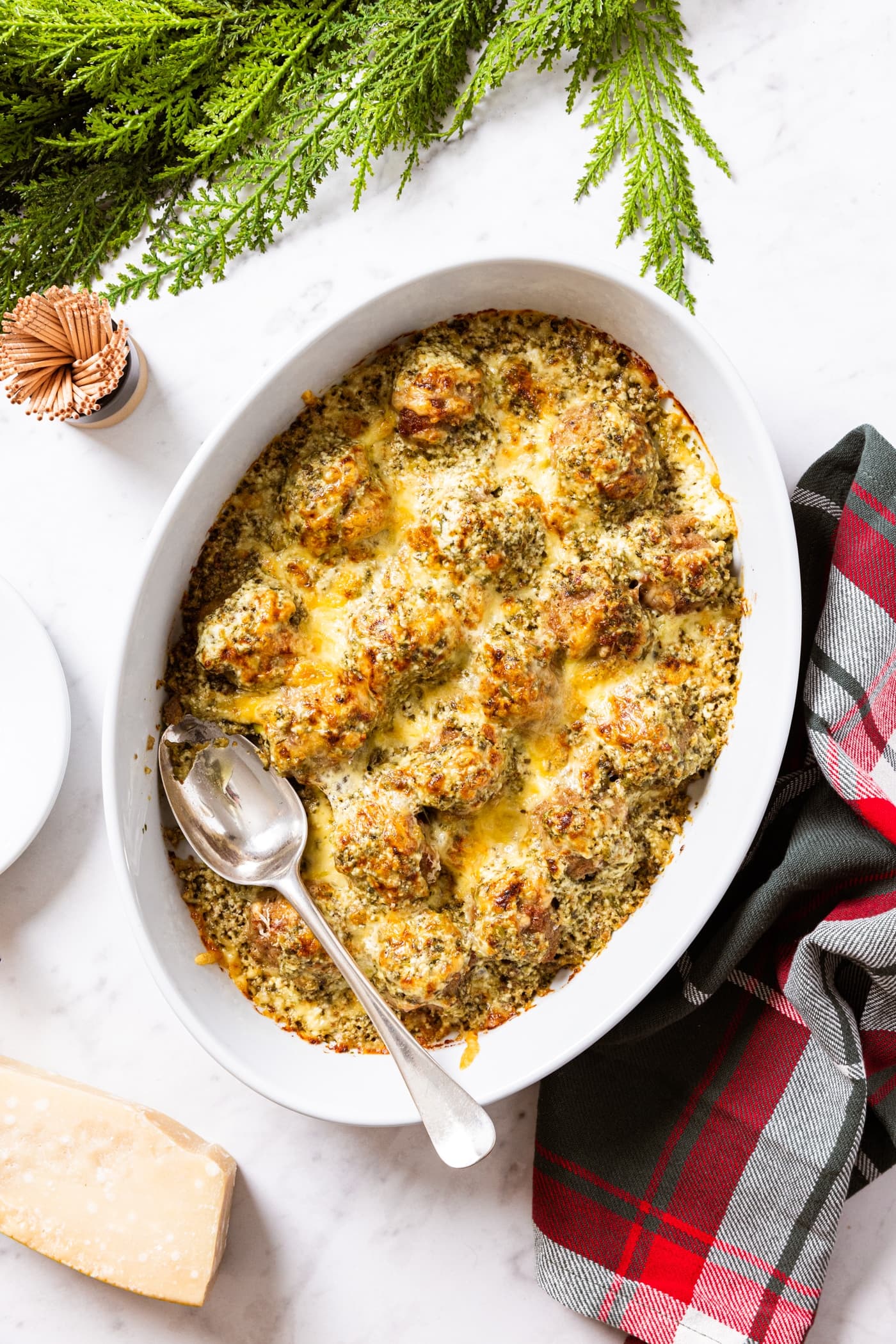 Oval white baking dish filled with baked meatballs sprinkled with pesto and Parmesan cheese sitting on marble surface with red and green Christmas napkin with greenery along the top.