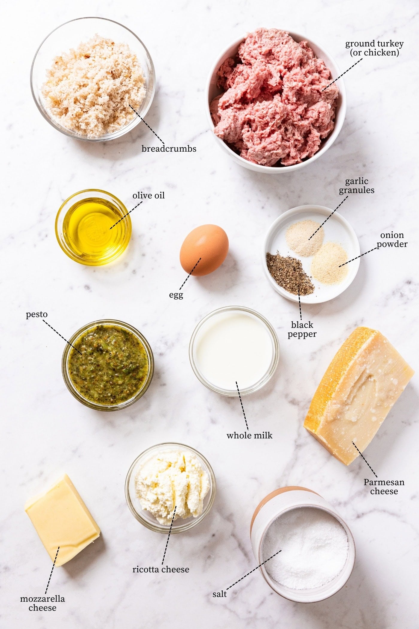 White marble surface filled with all ingredients needed to make pesto meatballs including raw ground meat, breadcrumbs, Parmesan cheese, pesto sauce, olive oil, egg, and more.