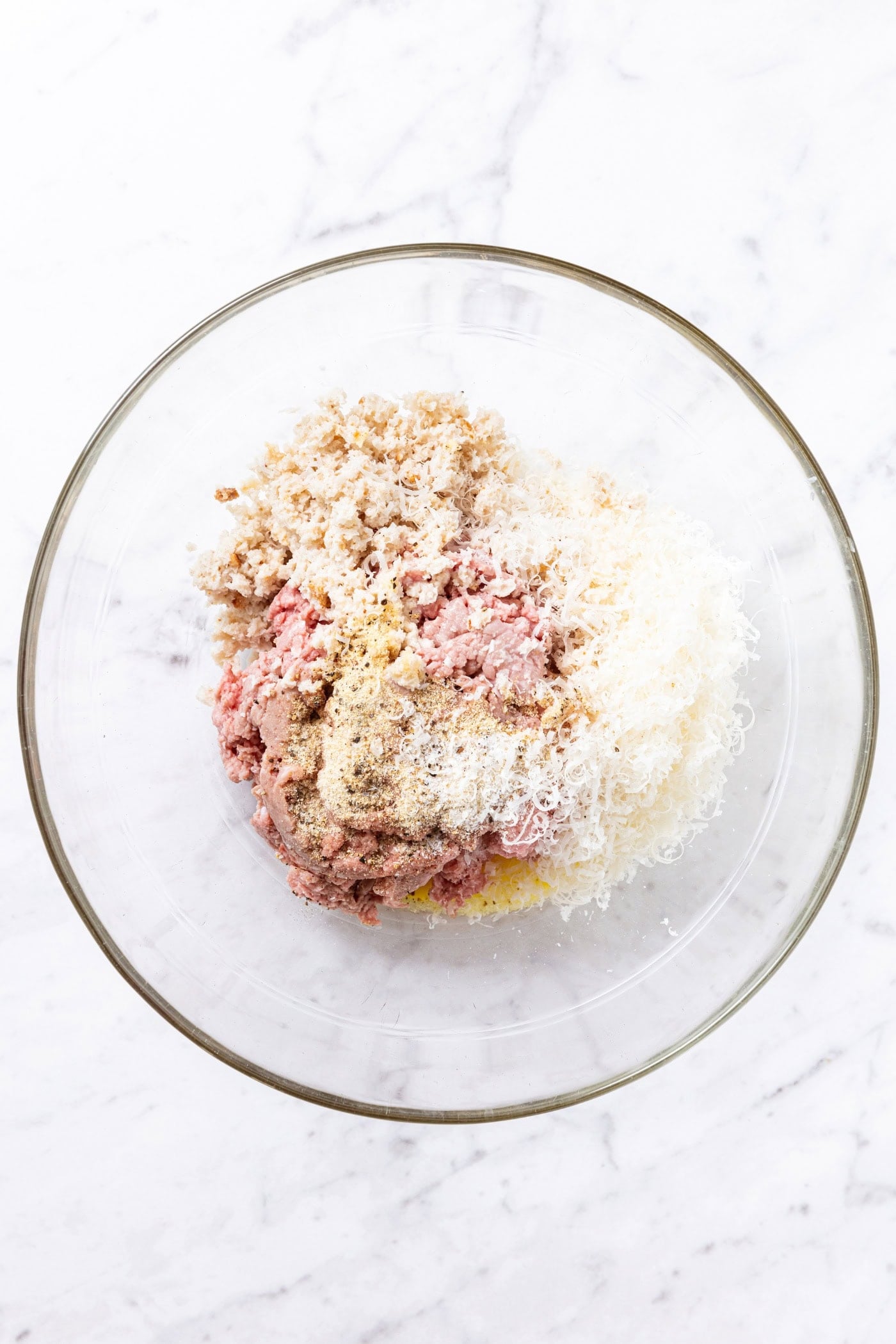 Glass bowl filled with all ingredients needed to make meatballs including ground meat, breadcrumbs, and cheese all on white marble surface.