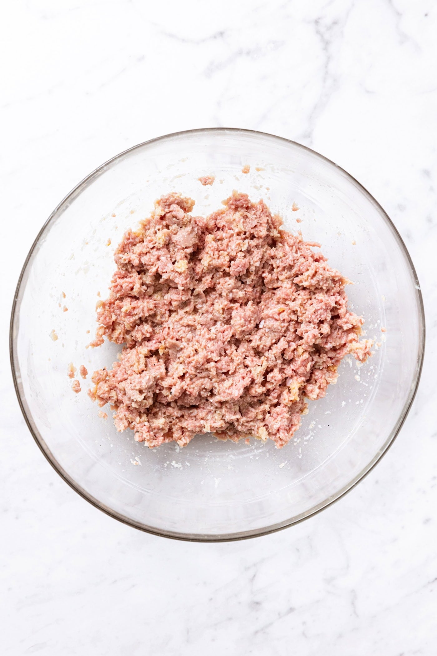 Glass bowl sitting on white marble with all ingredients mixed together for meatballs looking like ground meat.